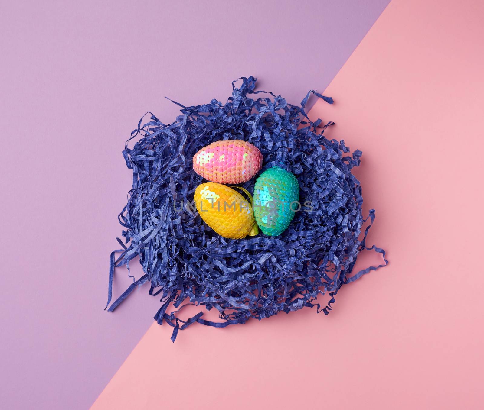 decorative Easter eggs from sequins lie in a nest of strips of paper on a colored background, top view