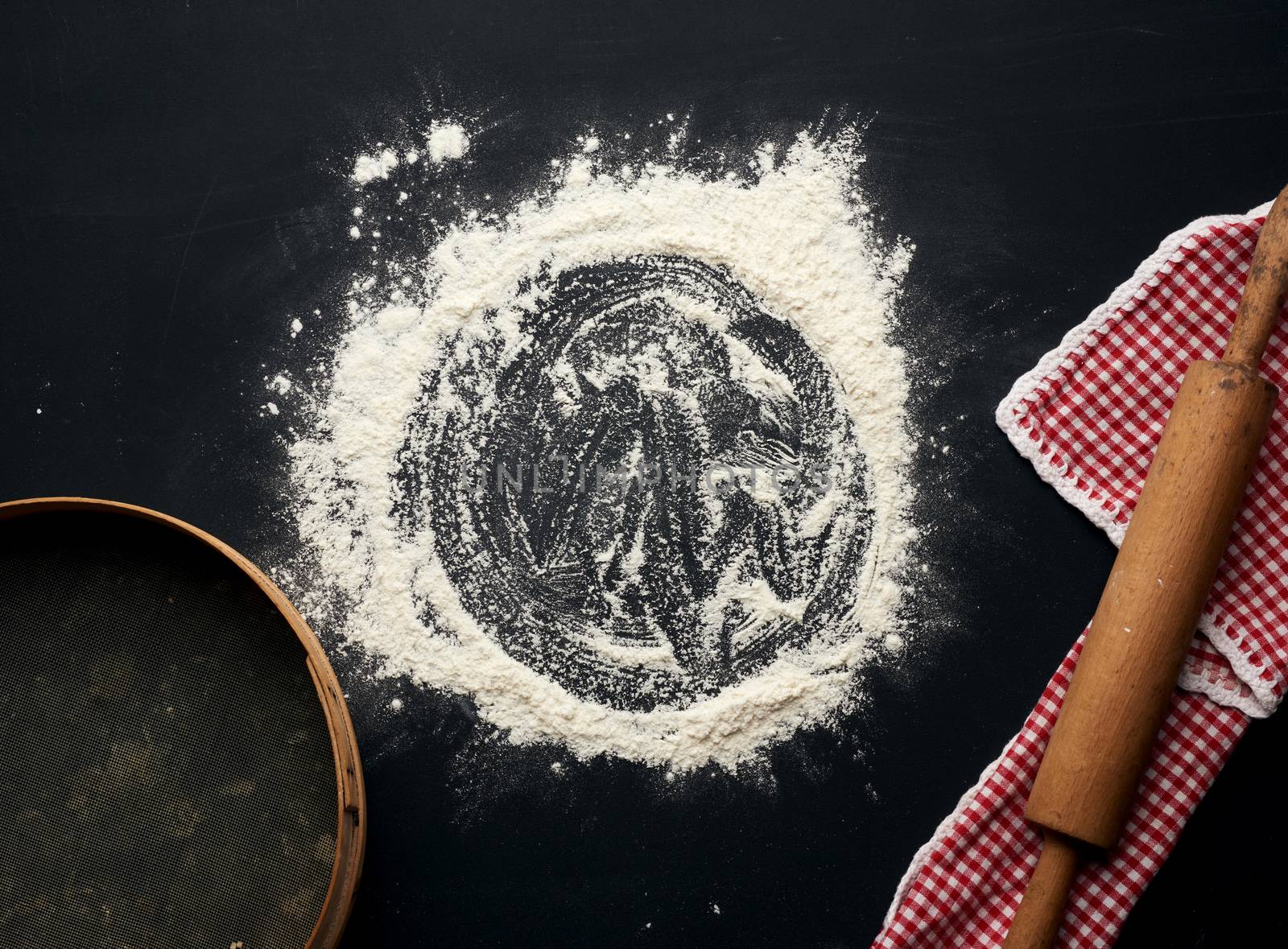 white wheat flour scattered on a black table and a wooden rolling pin on a black table, top view