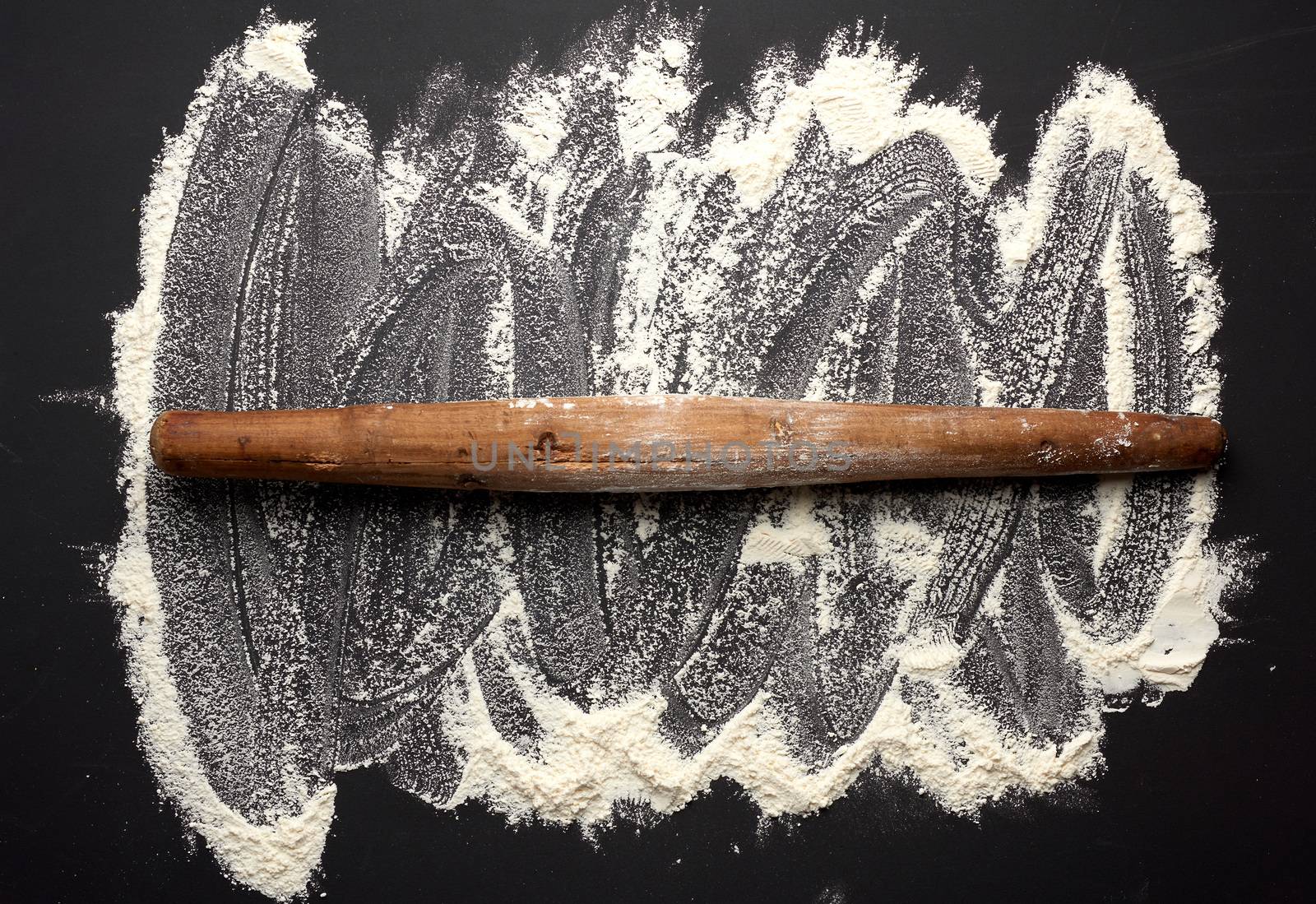 white wheat flour scattered on a black table and very old brown wooden rolling pin on a black table, top view