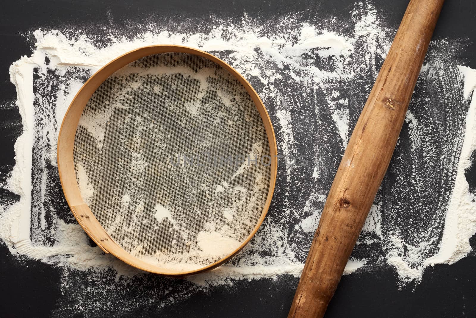 white wheat flour scattered on a black table and very old brown wooden rolling pin, round sieve, top view