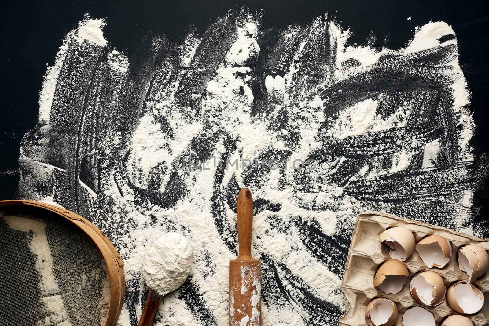sprinkled white wheat flour, wooden rolling pin, round sieve and chicken eggshell in a paper tray, top view, kitchen backdrop