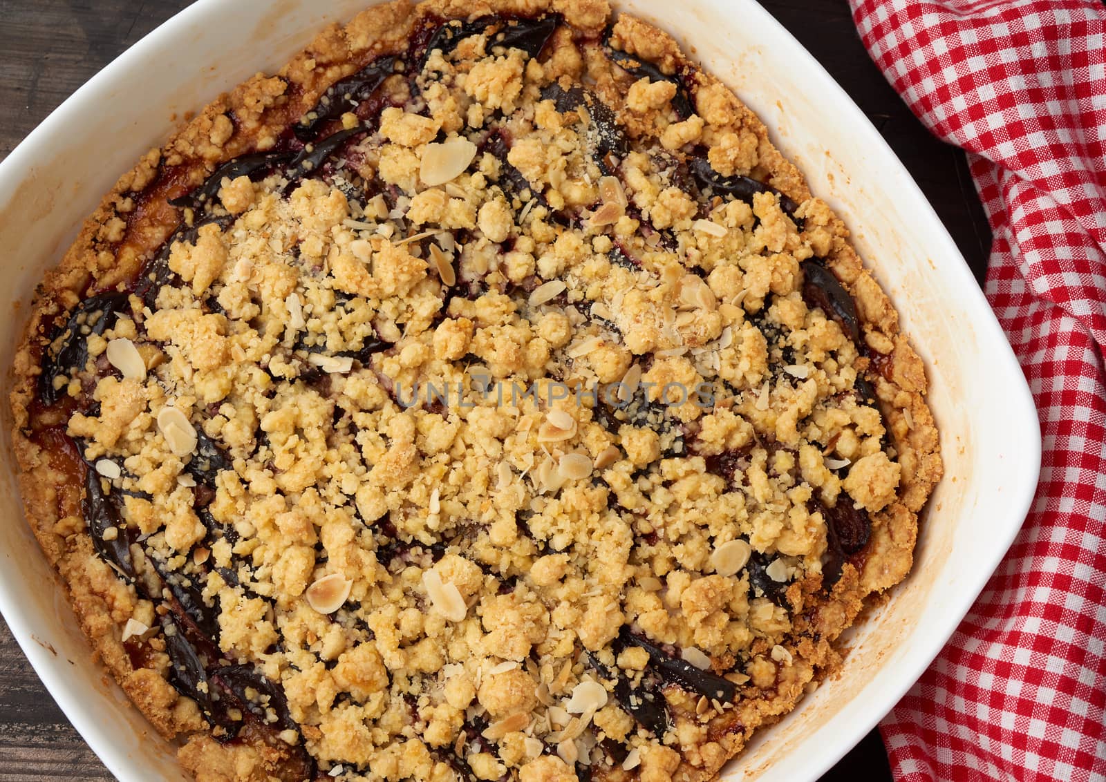baked crumble pie with blue plum in a white ceramic plate on a wooden table, tasty dessert, top view