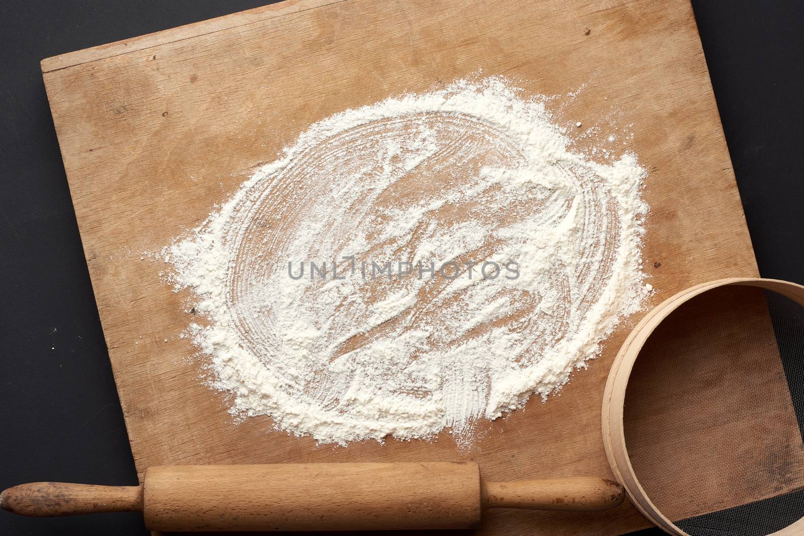 white wheat flour scattered and a wooden rolling pin, round sieve on a black table, top view