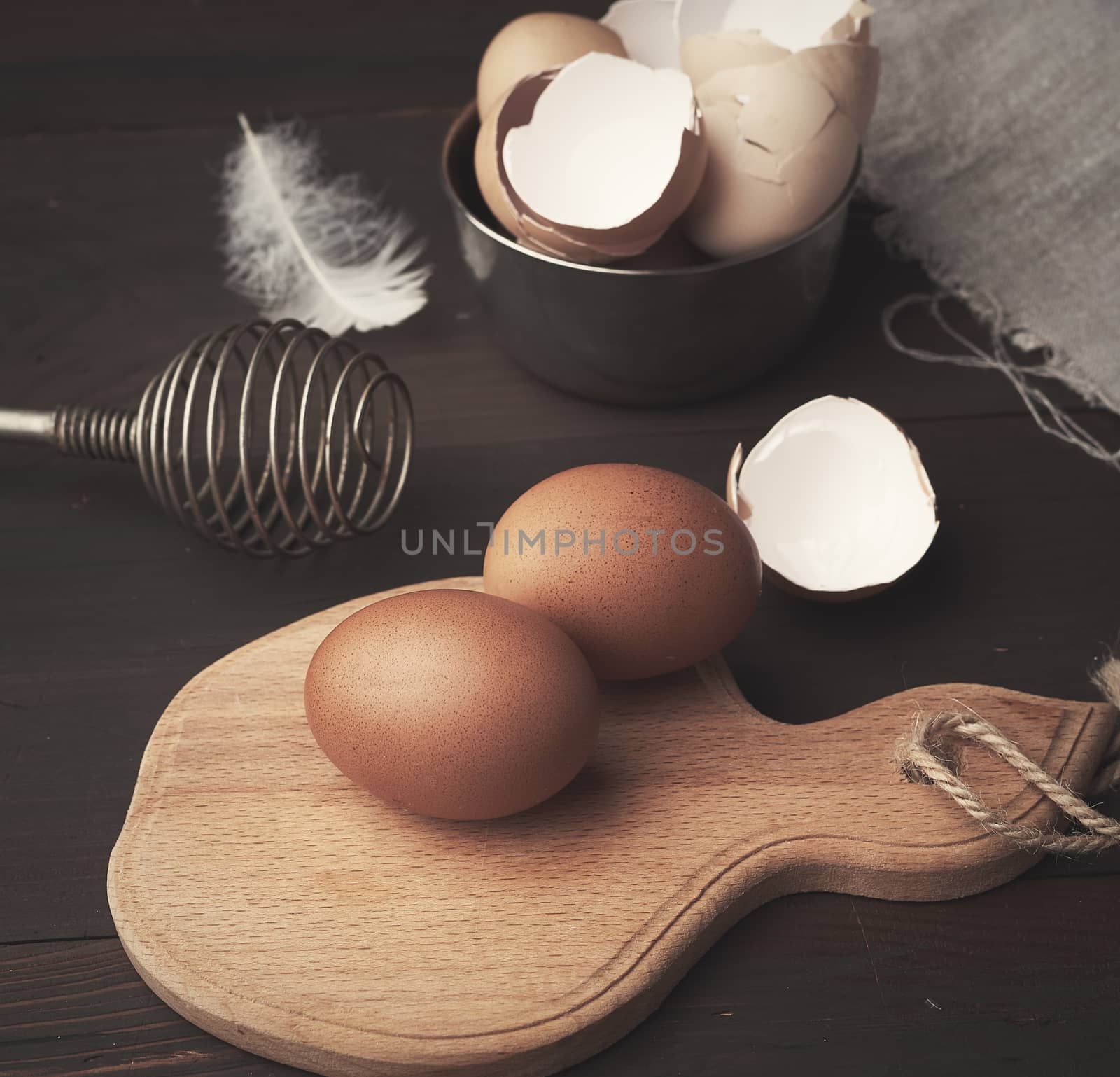 brown whole chicken eggs on a wooden board, behind a whisk for whipping and a shell