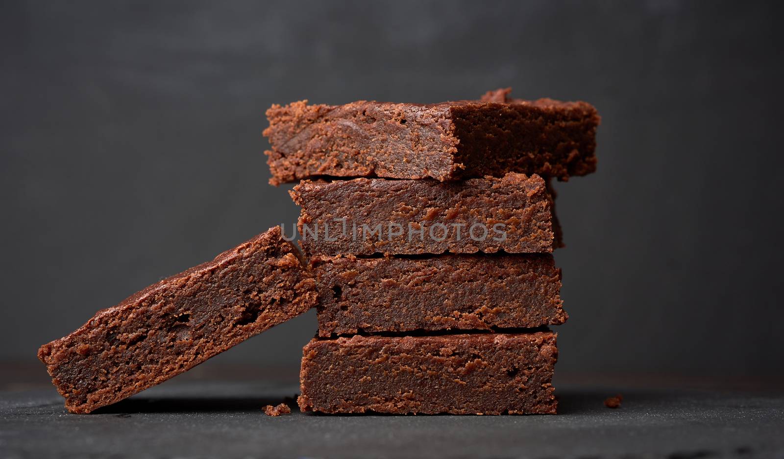 stack of square baked pieces of brownie chocolate cake on a black background, delicious dessert