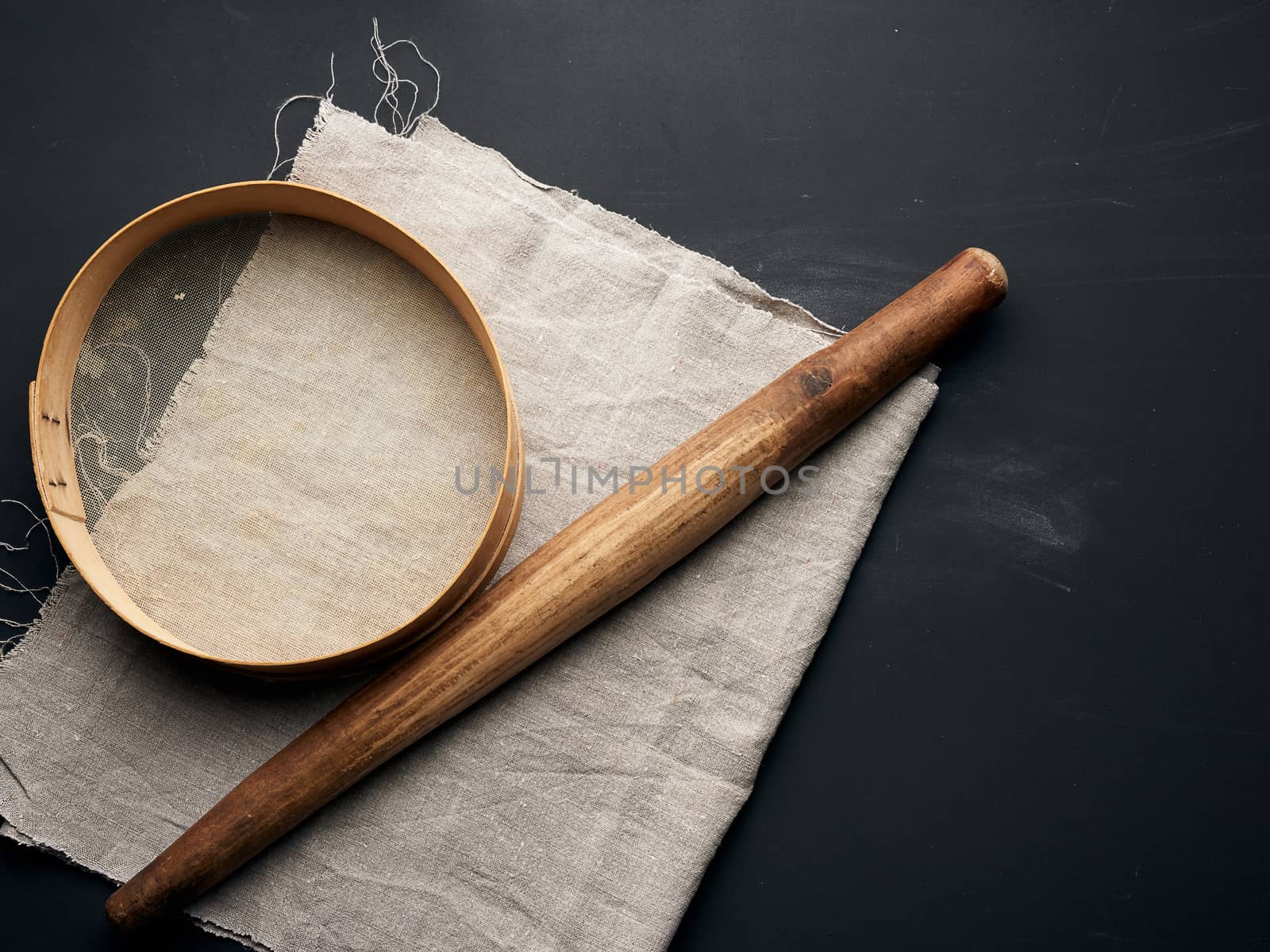 vintage wooden rolling pin and a round sieve lie on a gray linen napkin, black table, top view