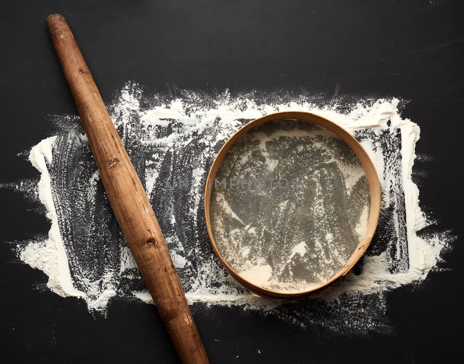 white wheat flour scattered on a black table and very old brown wooden rolling pin, round sieve, top view