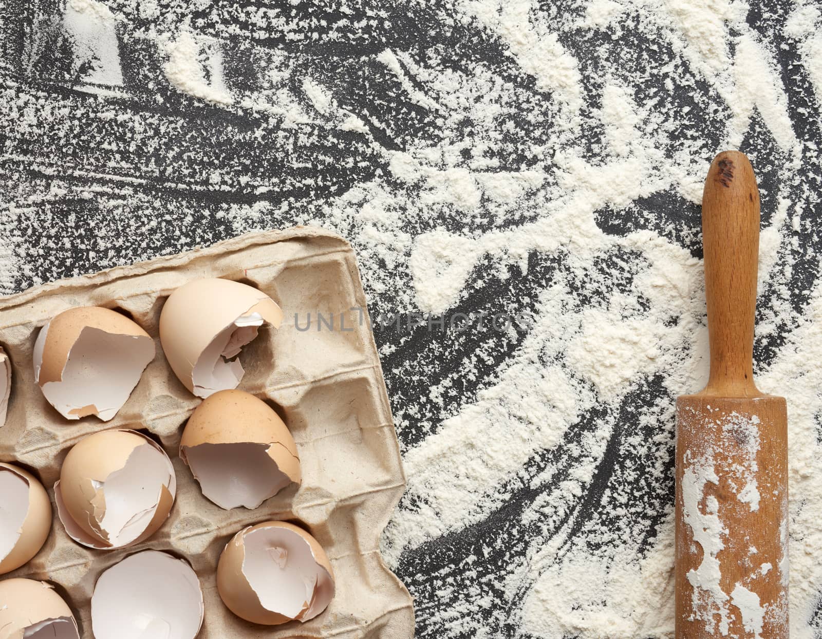 sprinkled white wheat flour, wooden rolling pin and chicken eggshell in a paper tray, top view, kitchen backdrop