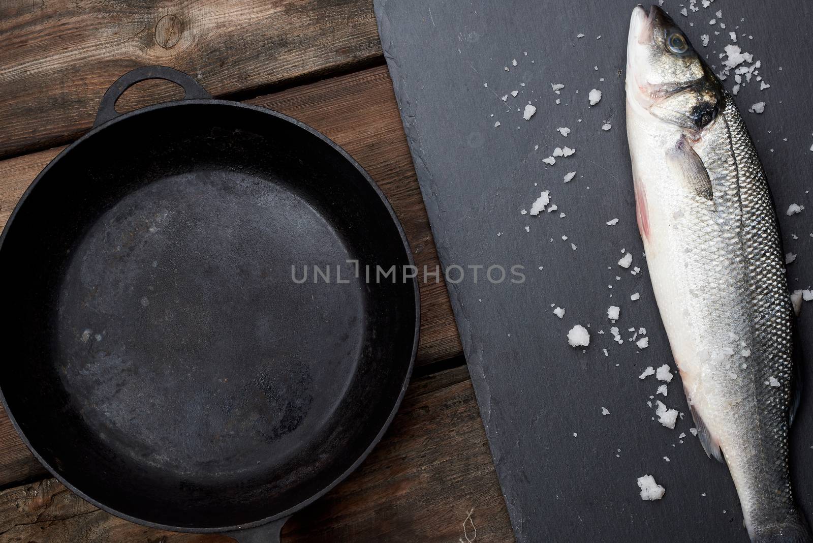 fresh whole sea bass fish on a black board, next to it is an empty round black frying pan on a wooden table, top view