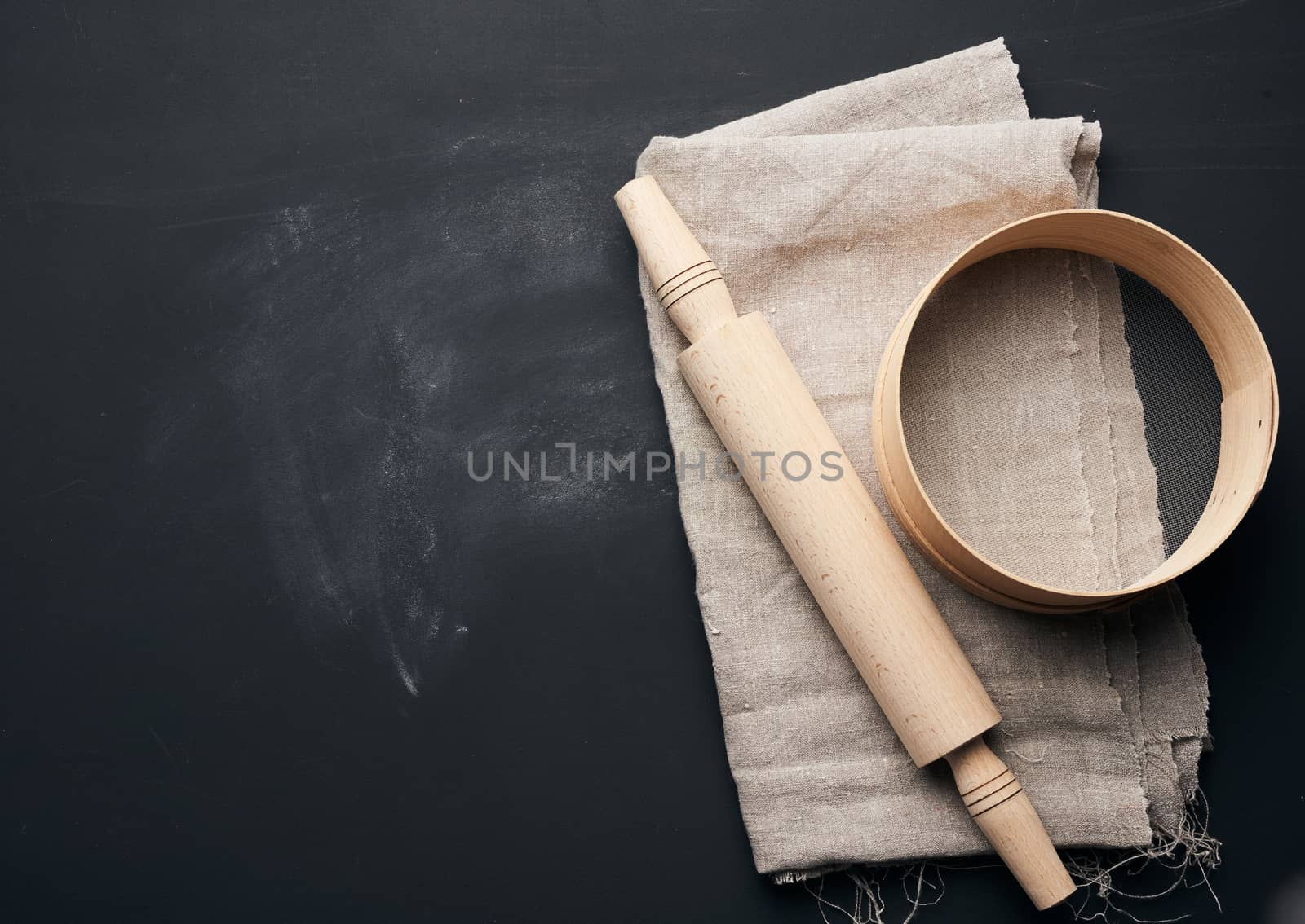 wooden rolling pin and a round sieve lie on a gray linen napkin, black table, top view, copy space