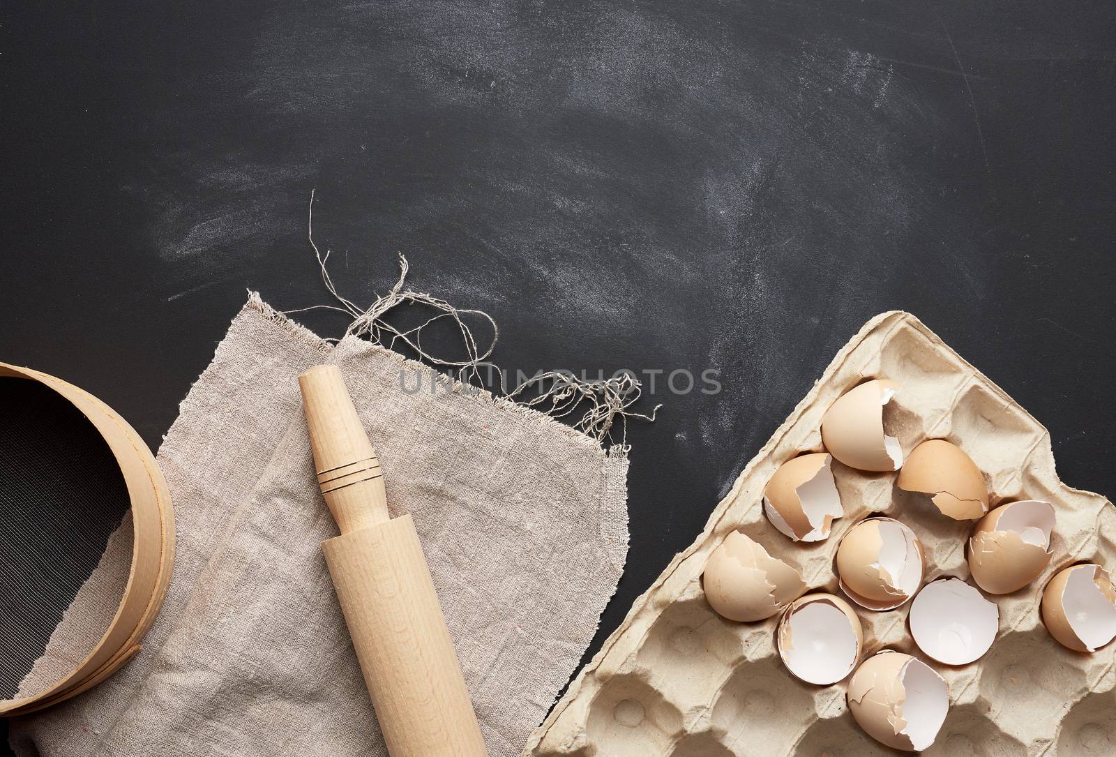 wooden round sieve, tray of chicken eggs and rolling pin on a black table, ingredients for kneading dough, top view, copy space