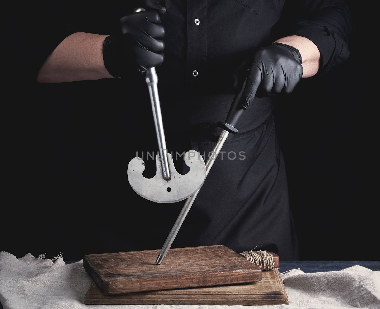 male cook in black uniform and black latex gloves holds a large sharp vintage knife for meat and vegetables and a metal sharpener for knives, black background