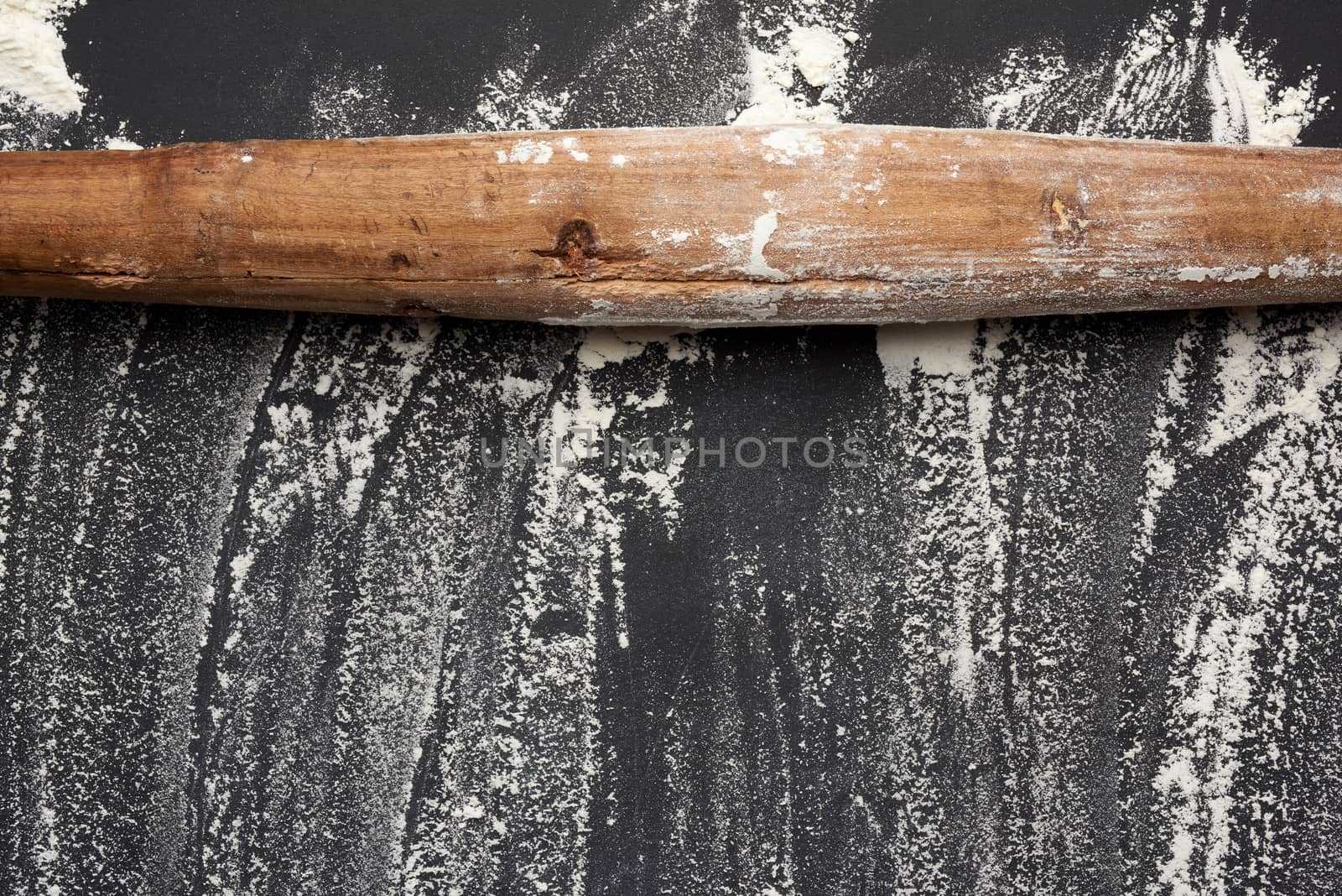 white wheat flour scattered on a black table and very old brown wooden rolling pin on a black table, top view