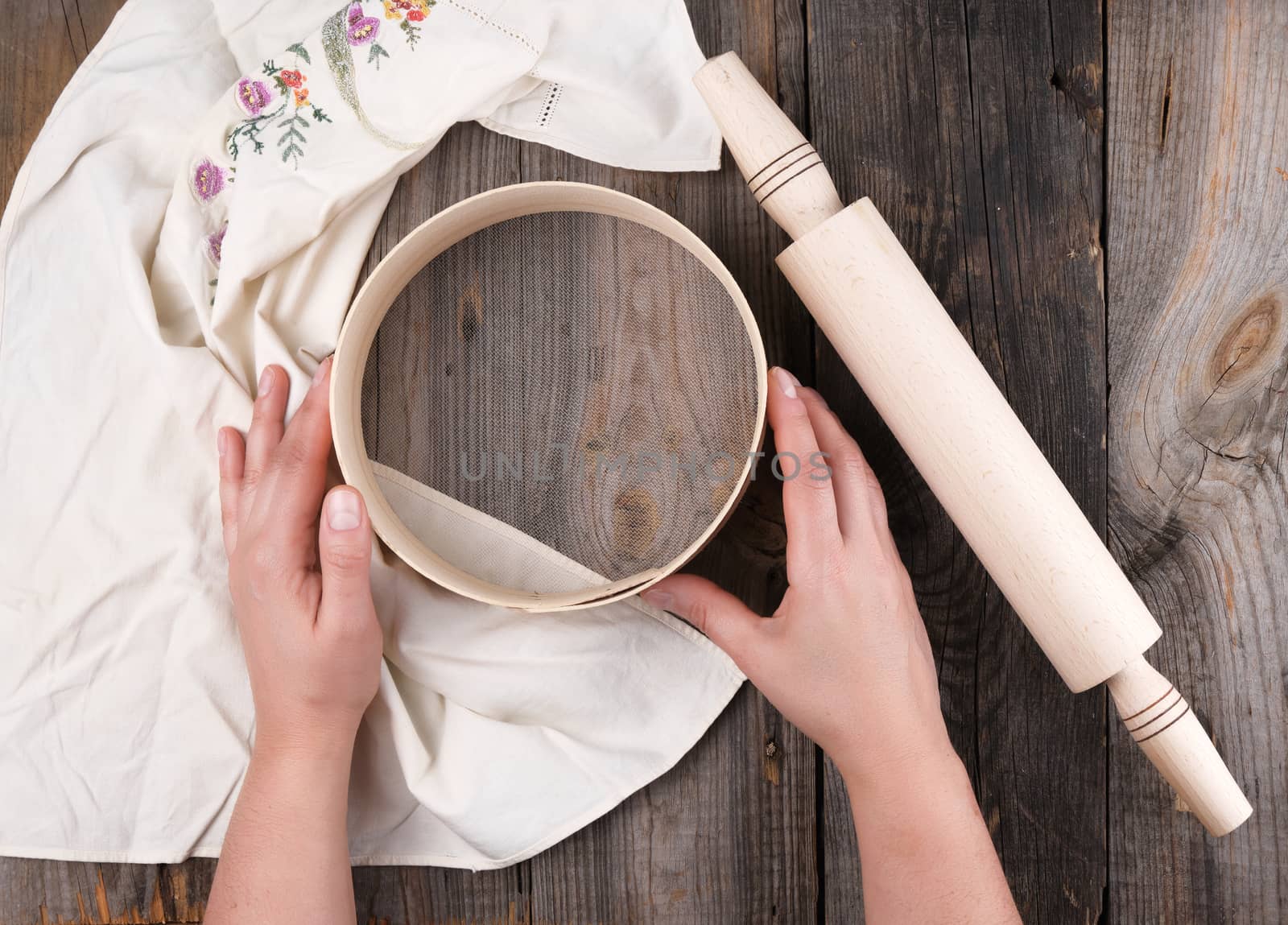 female hands holding a round wooden sieve for flour, next to an wooden rolling pin on a gray table