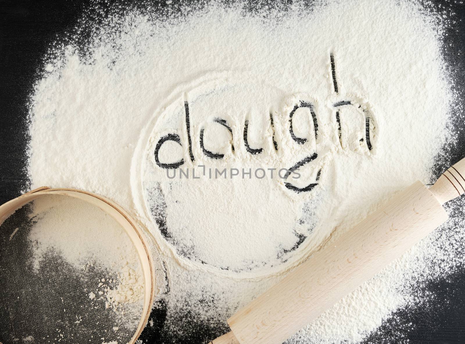 sprinkled white wheat flour on a black table and the inscription dough, next to a wooden round sieve and rolling pin, top view