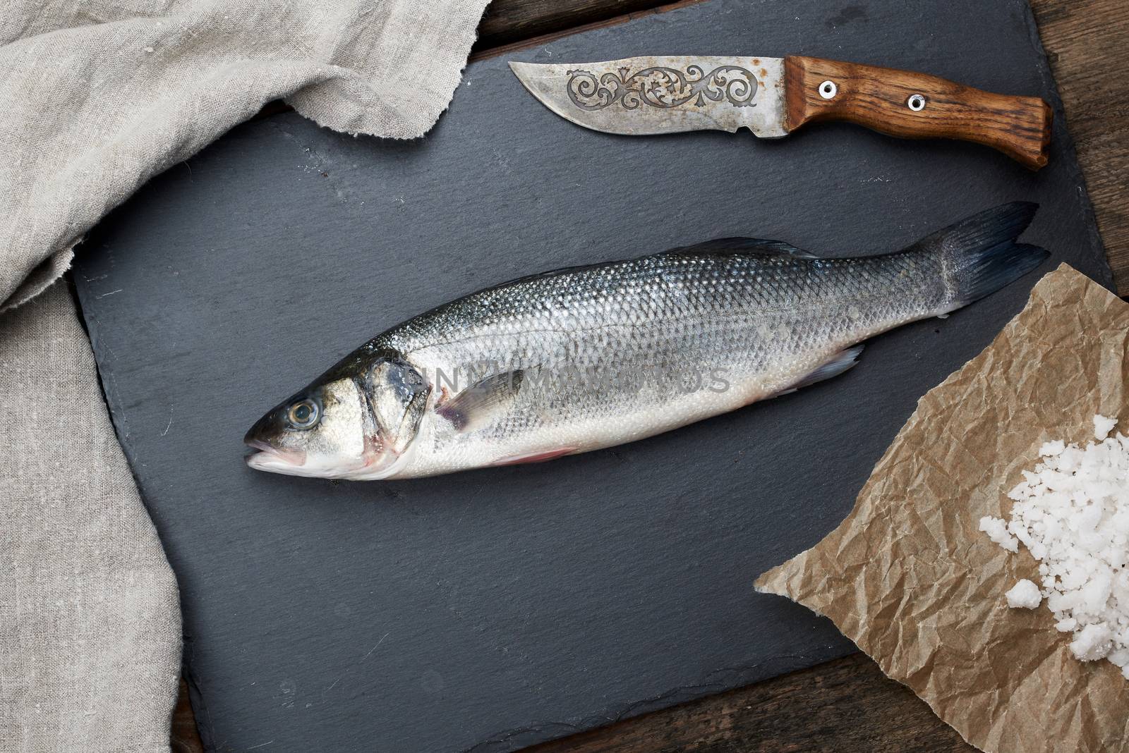 fresh whole sea bass fish on a black board, old knife and white salt, top view