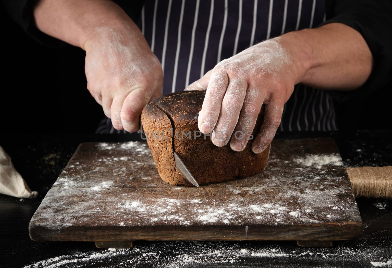 chef in black uniform holds a kitchen knife in his hand and cuts off pieces of bread from a baked brown rye flour loaf on a wooden board