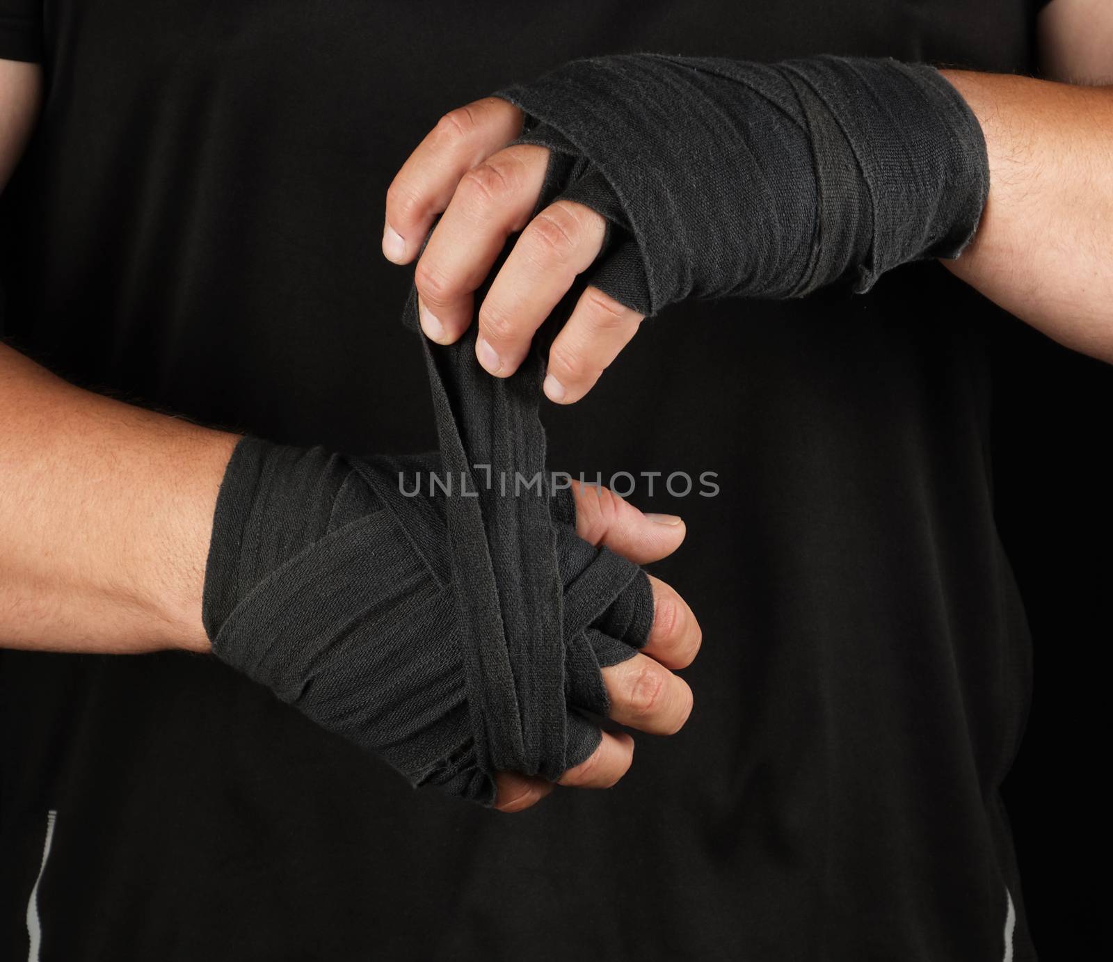 adult athlete stand in black clothes and wrap his hands in black textile elastic bandage before training, black background, muscular body