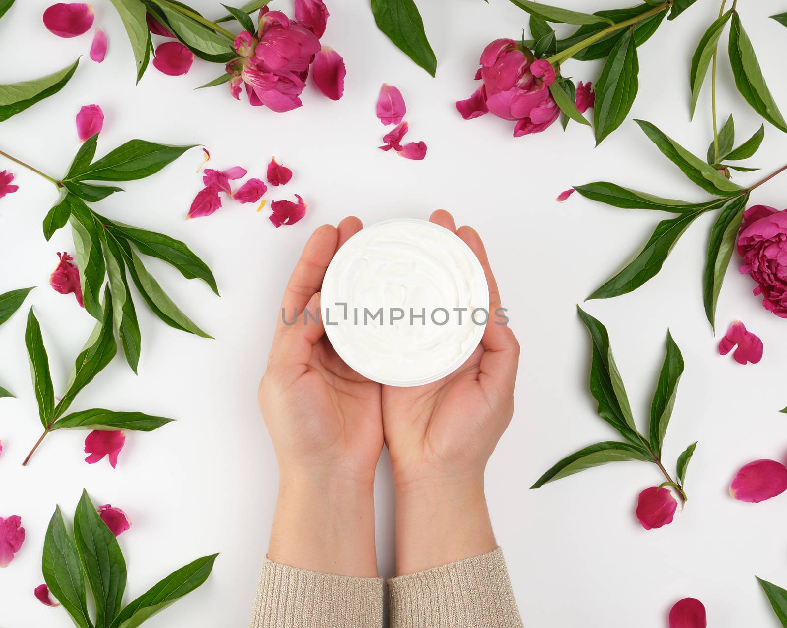 two female hands and a jar with thick cream and burgundy flowering peonies with green leaves, top view, concept of anti-aging procedures for rejuvenating and moisturizing the skin of hands