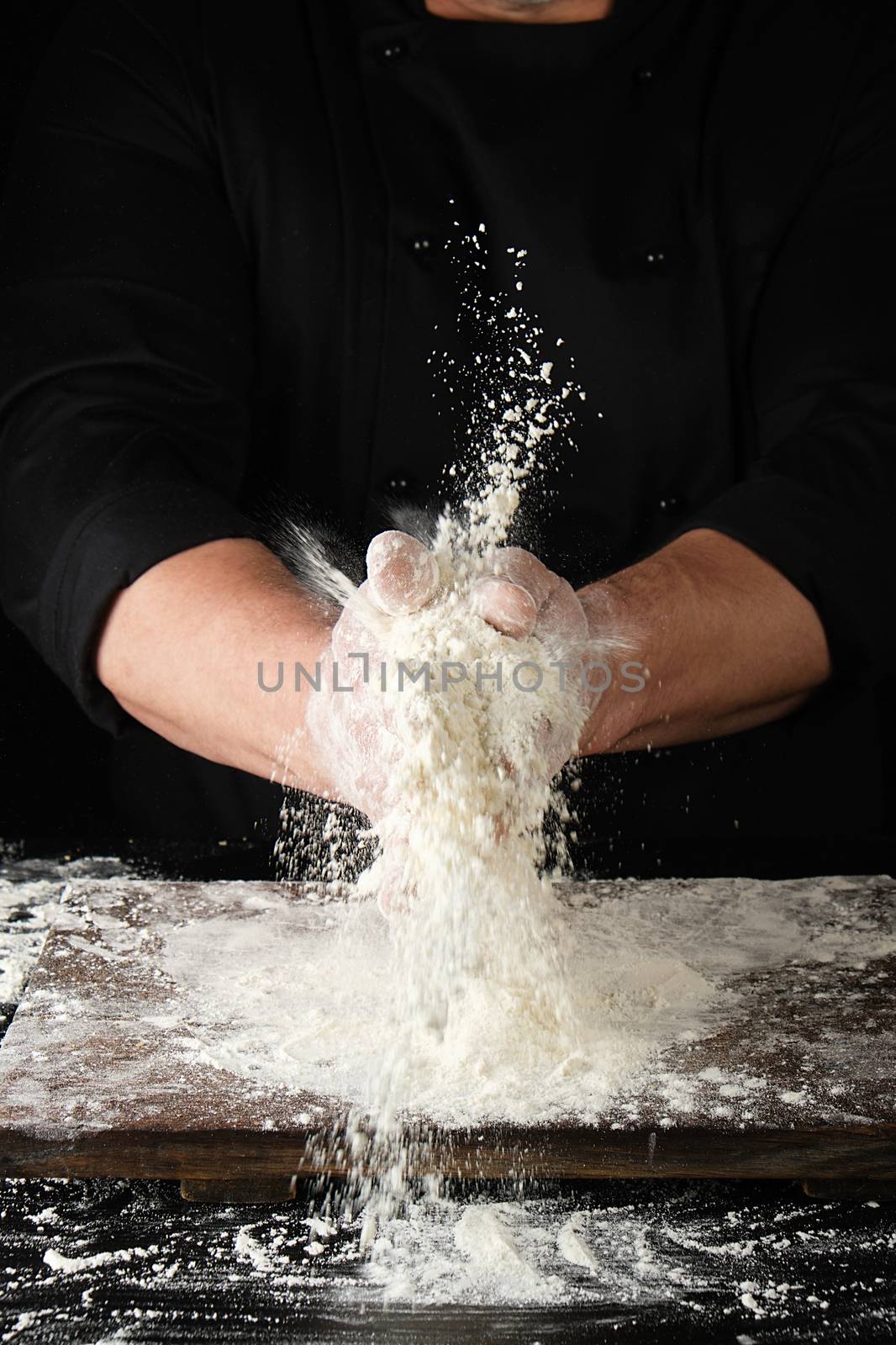 chef in black uniform sprinkles white wheat flour in different directions, product scatters dust, black background