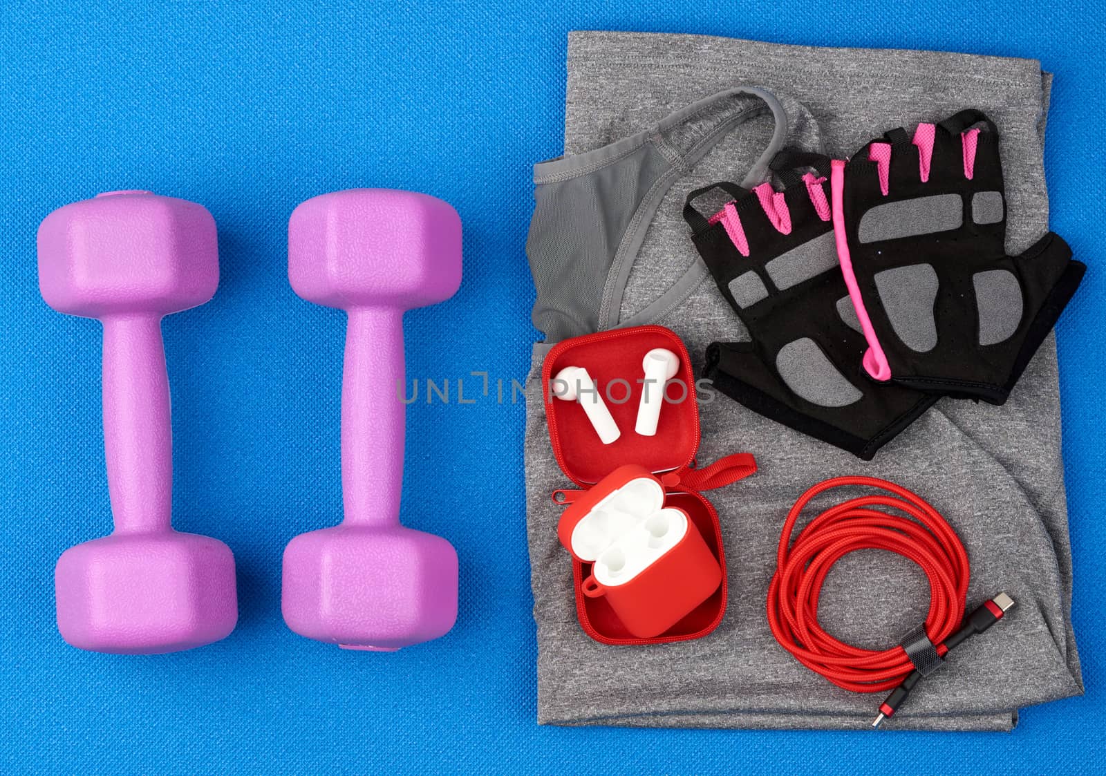 plastic purple dumbbells, gray sportswear, water and wireless headphones on a blue background from a sports mat, fitness kit, flat lay, copy space