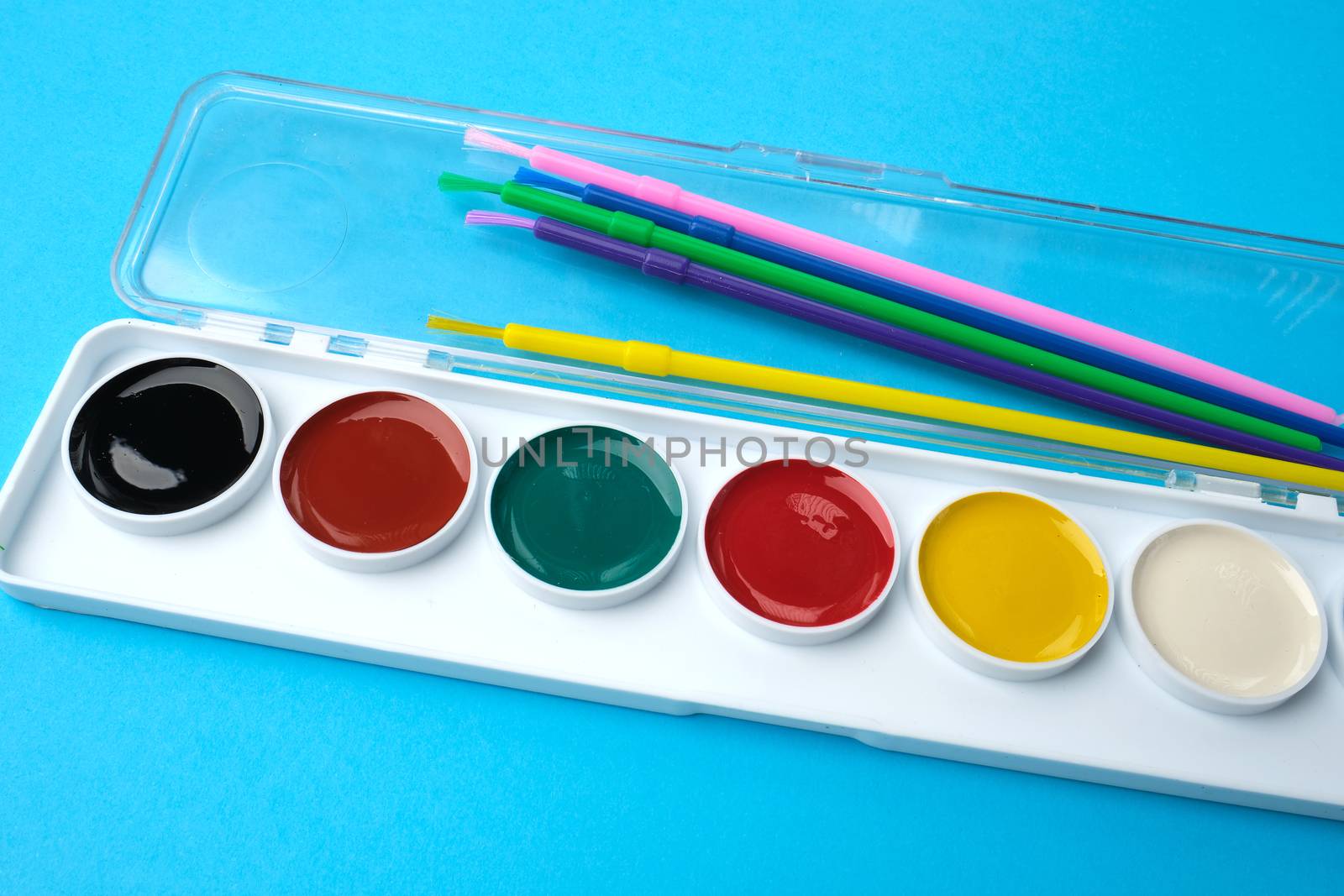 multicolored watercolor paints in a plastic box and brushes on a blue background, set for drawing, close up