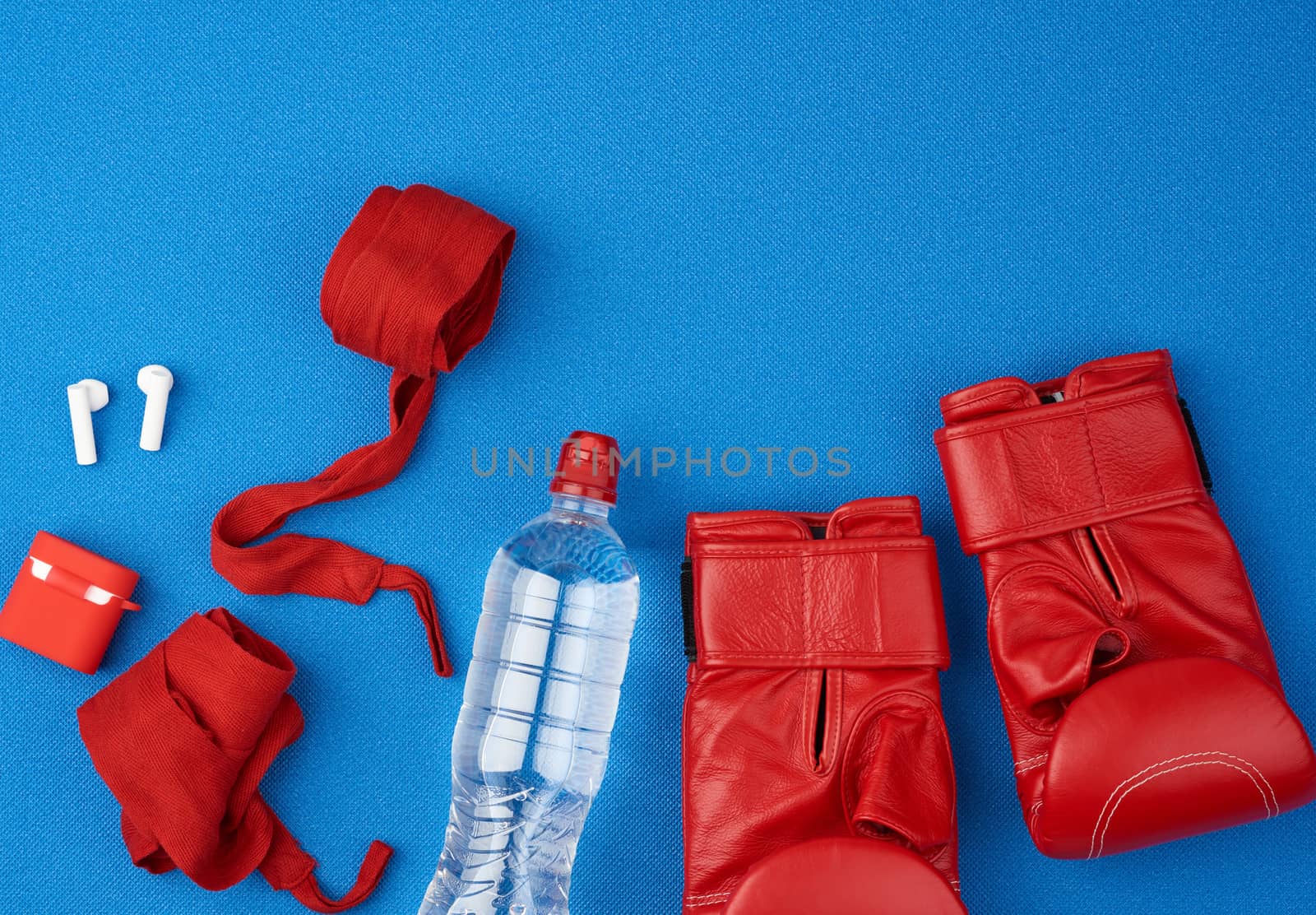 pair of red leather boxing gloves, a textile red elastic bandage for hands, wireless headphones and a bottle of water lie on a neoprene blue mat, sports kit