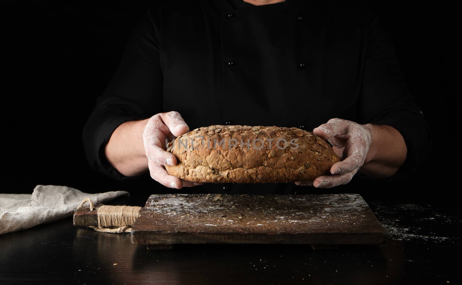 male hands are holding brown baked rye bread over wooden board with flour, black background