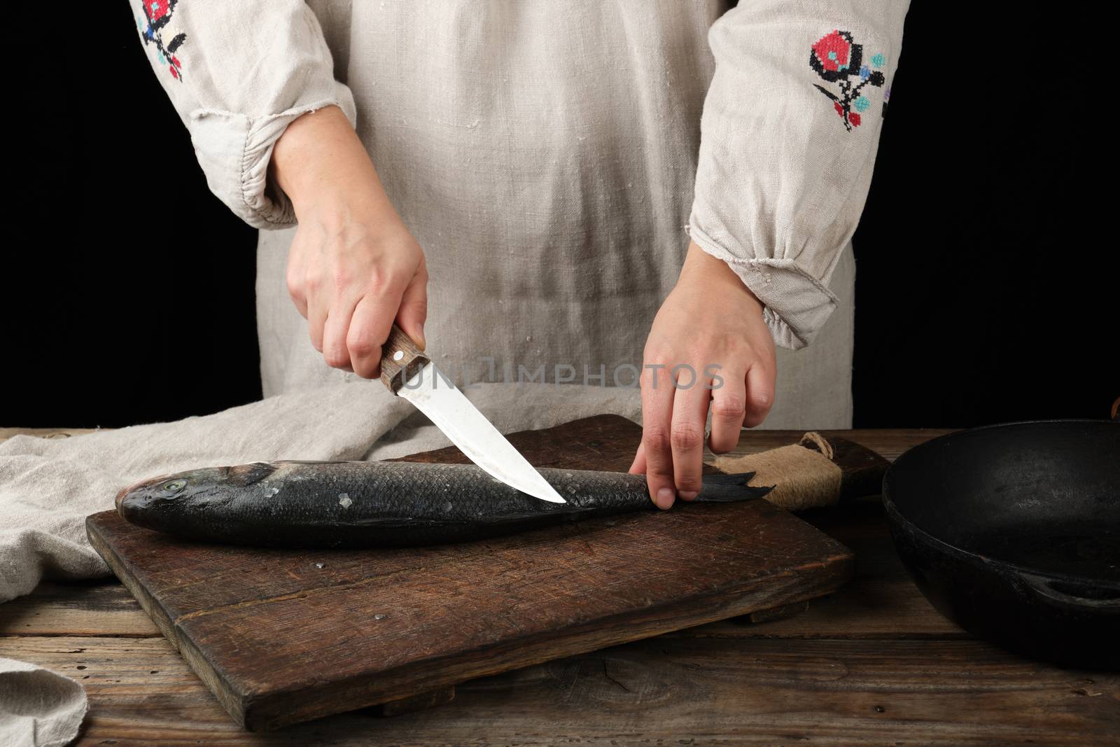 woman in gray linen clothes cleans the fish sea bass scales on a brown wooden board, black background