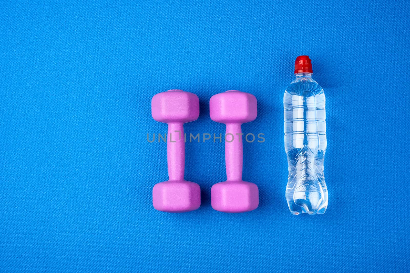 transparent plastic bottle of water and a pair of purple plastic dumbbells for sports and fitness on a blue mat, top view, copy space