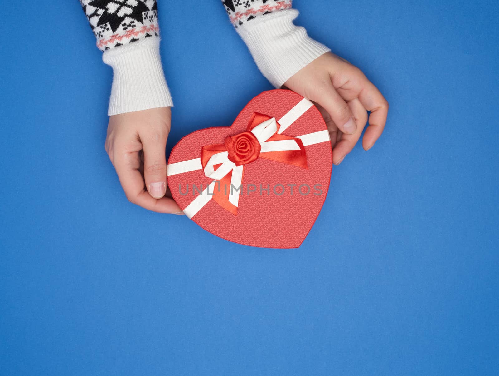 two female hands hold a paper closed red box in the shape of a heart on a dark blue background, festive backdrop, copy space