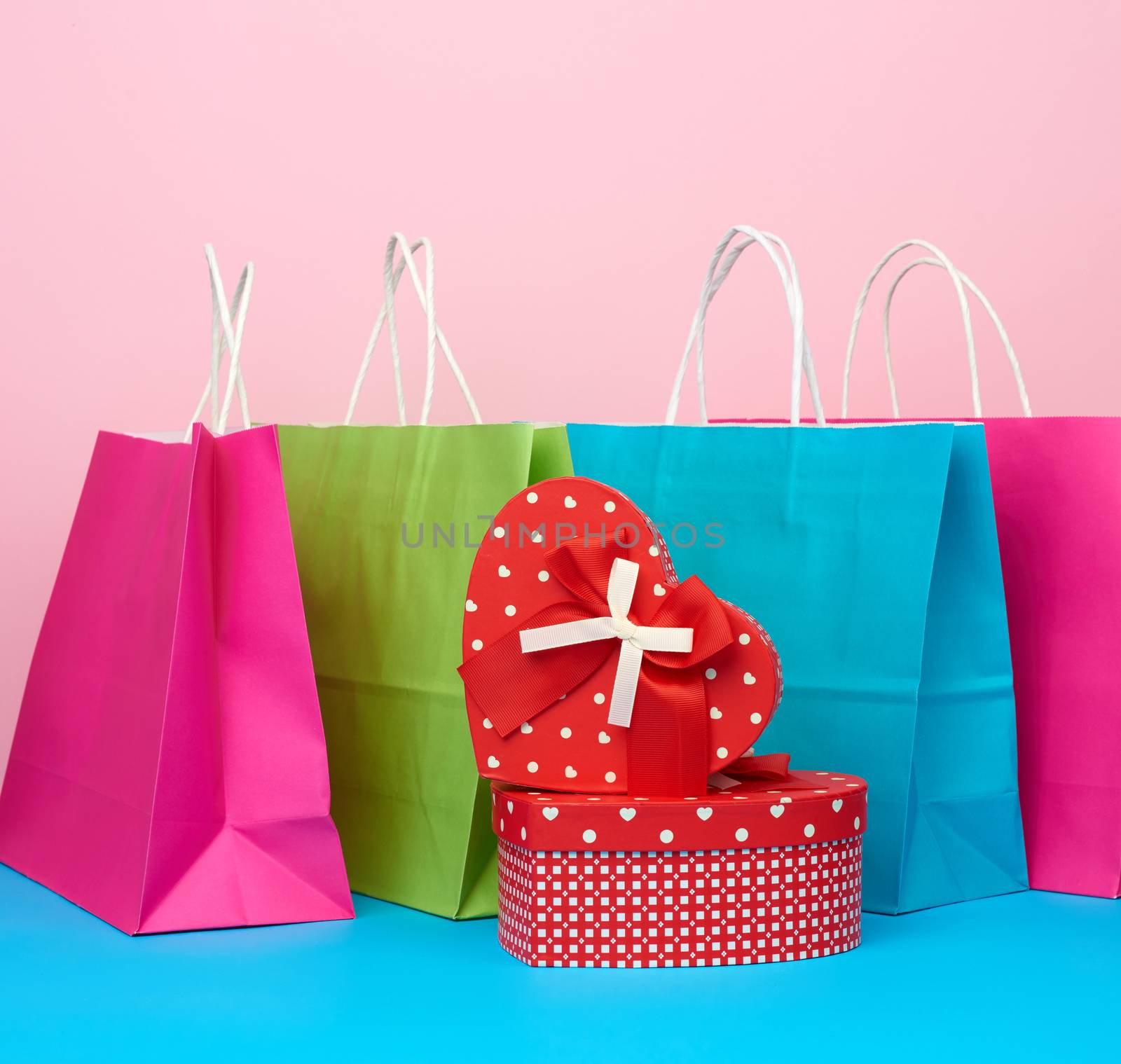 red paper box with a gift and paper bags for shopping stand on a pink-blue background