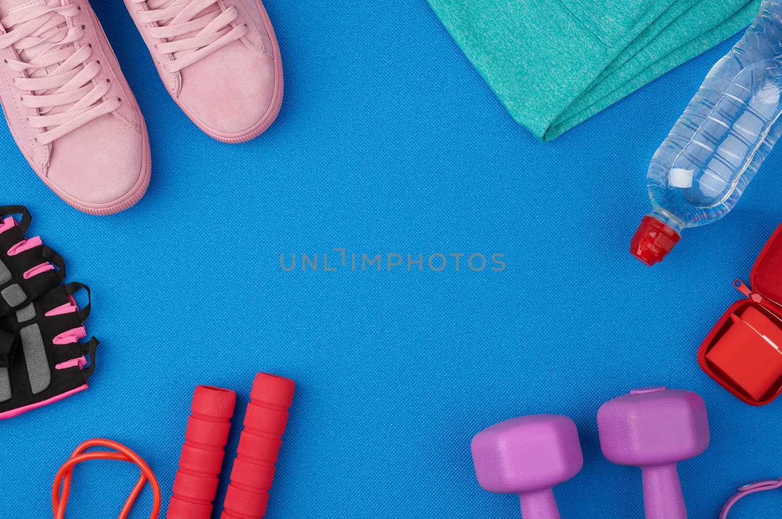 plastic purple dumbbells, sportswear, water , pink sneakers and wireless headphones on a blue background from a sports mat, fitness kit, flat lay, copy space