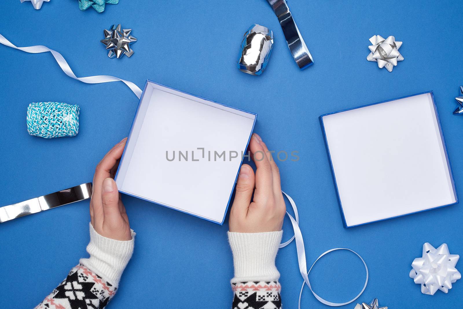 two female hands in a sweater hold a open square blue box with a bow on a blue classic background, top view, festive backdrop