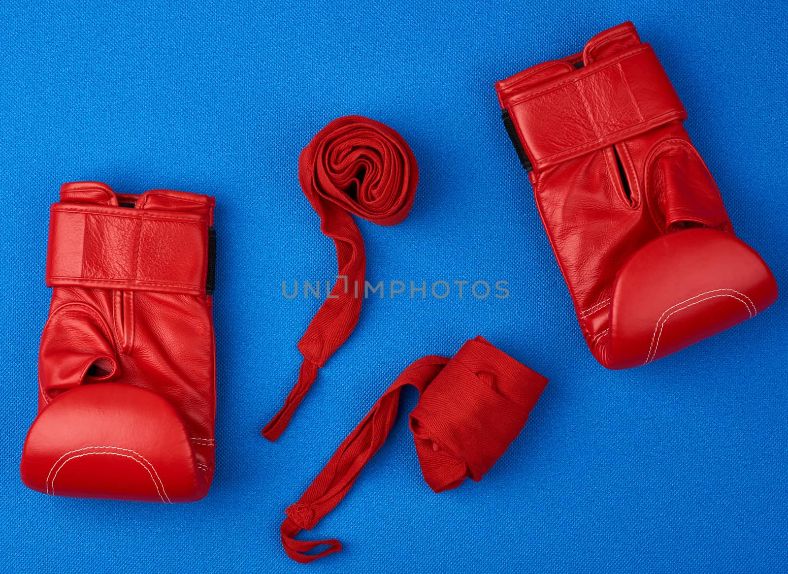 pair of red leather boxing gloves and a textile red elastic bandage for hands lie on a blue neoprene mat, sports set
