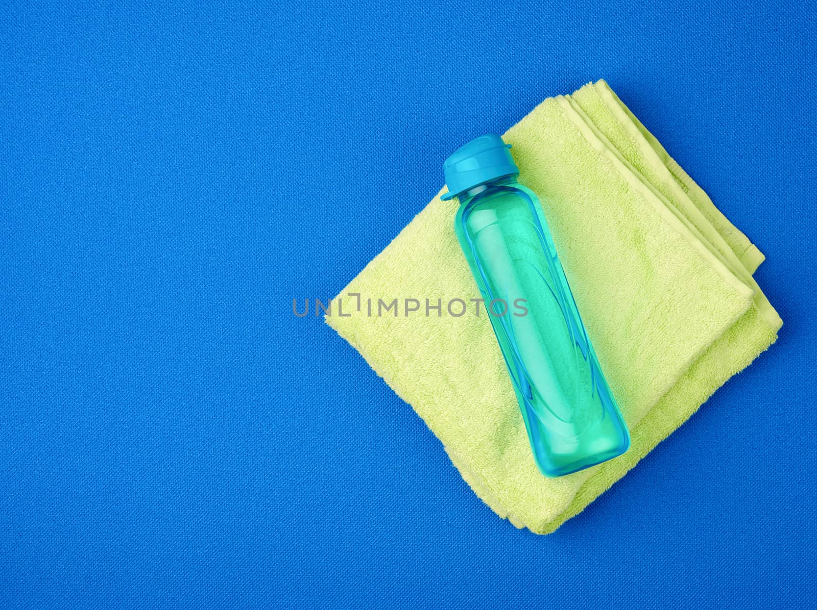 folded green terry towel and a transparent bottle of water on a blue background, concept of drinking more water while doing sports
