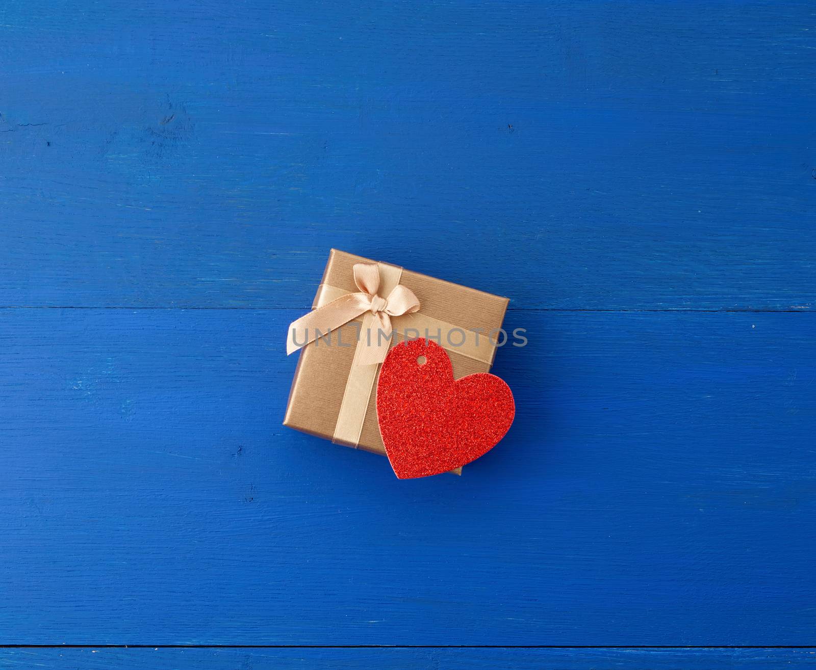 closed golden cardboard box with a gift and red paper heart, blue wooden background from boards, top view