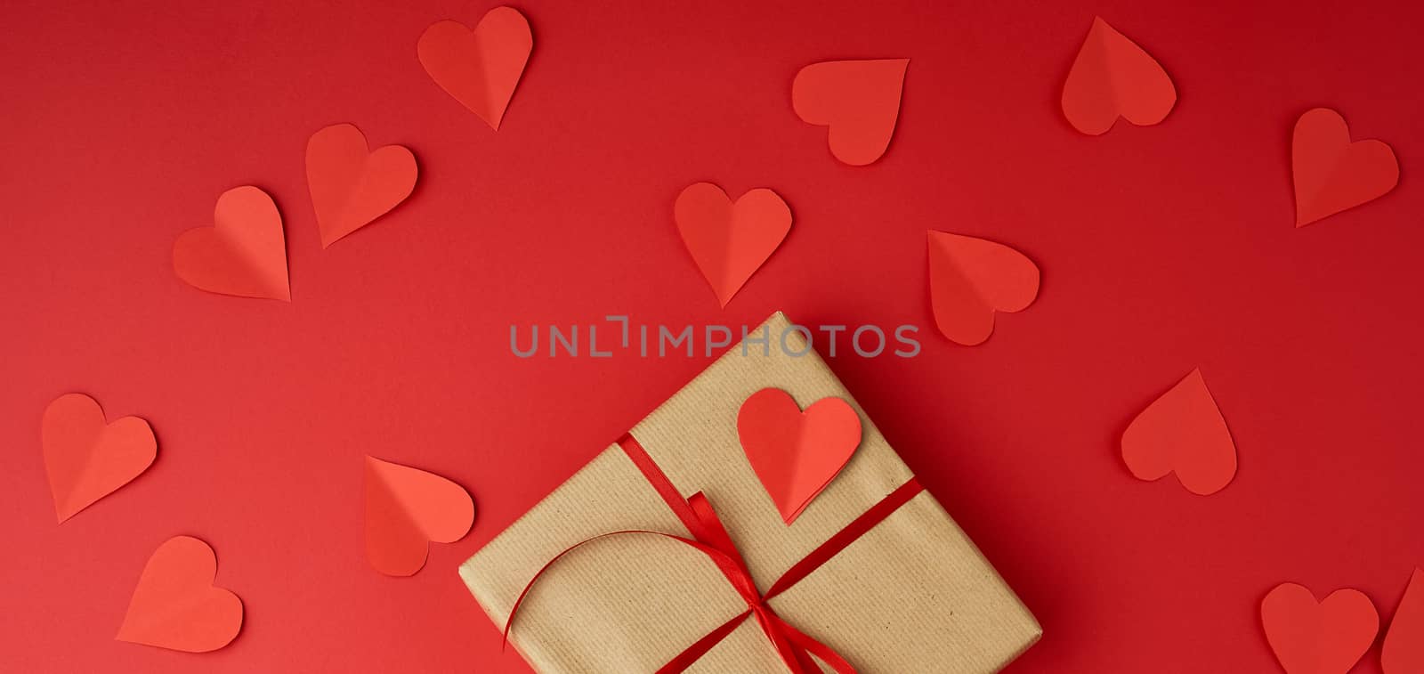 square box wrapped in brown kraft paper and tied with a red thin silk ribbon, cut out of paper hearts, red background, gift for Valentine's Day, birthday