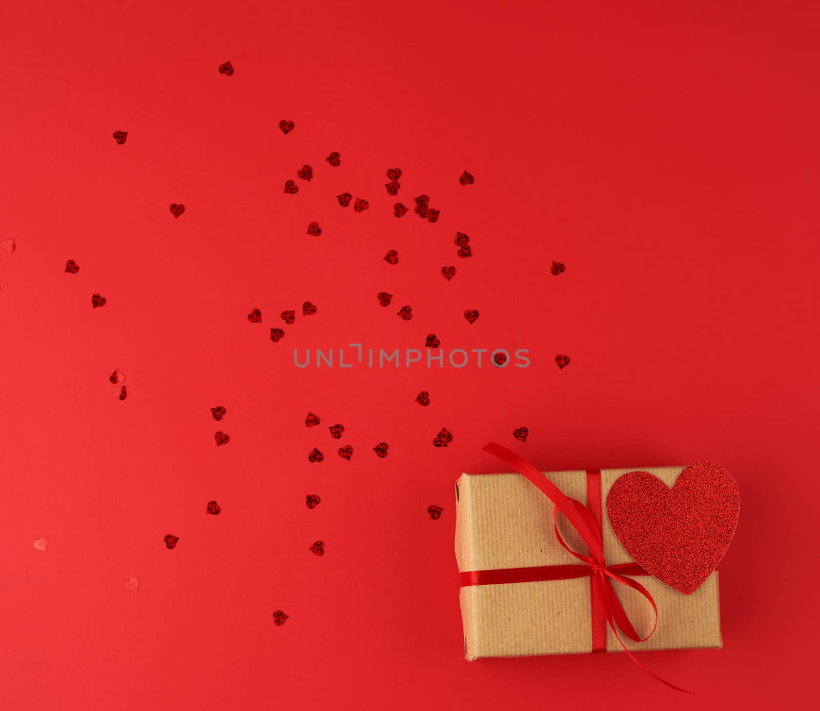 box wrapped in brown kraft paper and tied with a red thin silk ribbon, scattered confetti in the shape of hearts, red background, festive backdrop for Valentine's Day