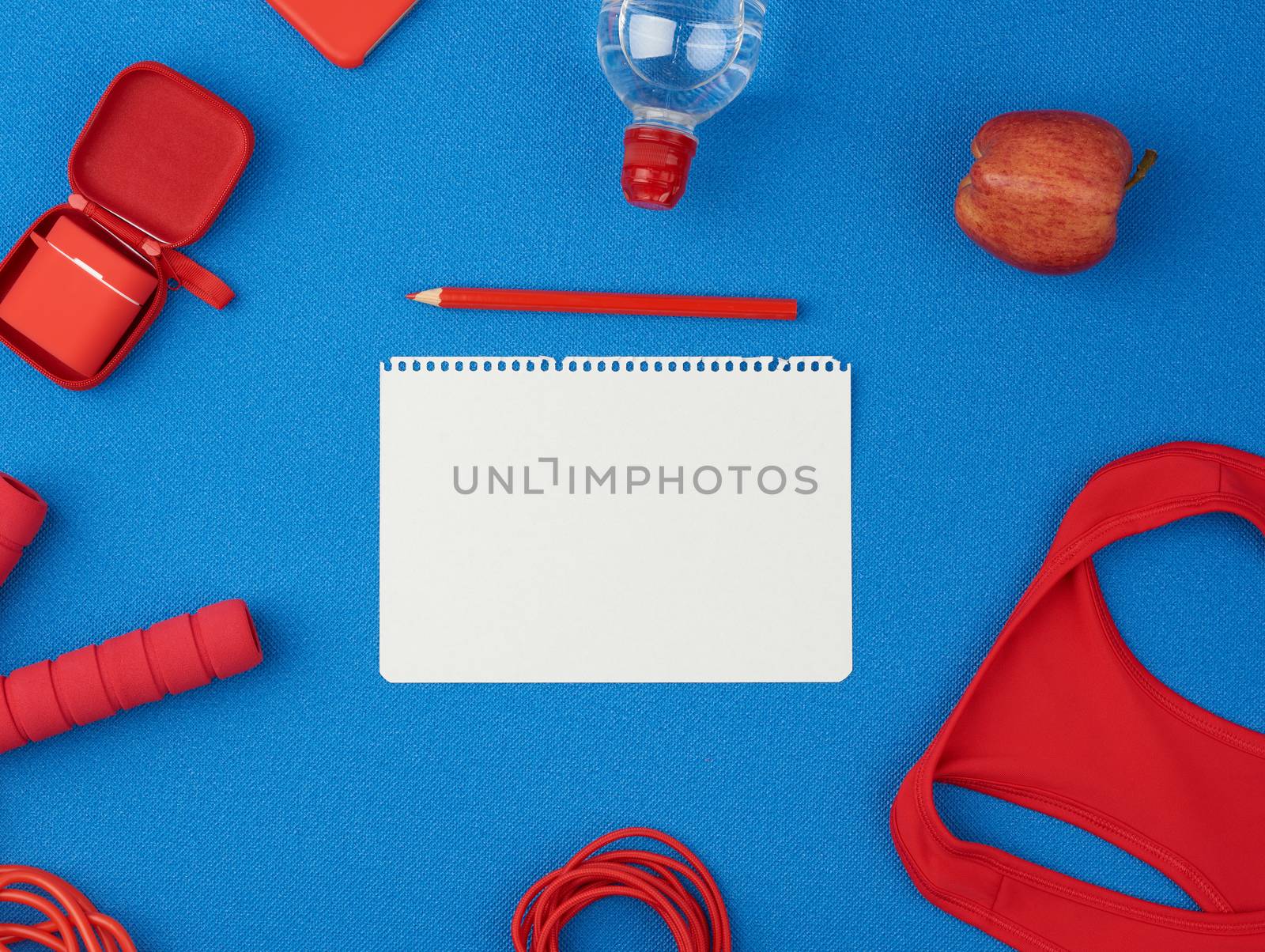 red sports shirt, red jump rope and wireless headphones in a red case on a blue background, top view, in the middle of an empty white sheet of paper, flat lay