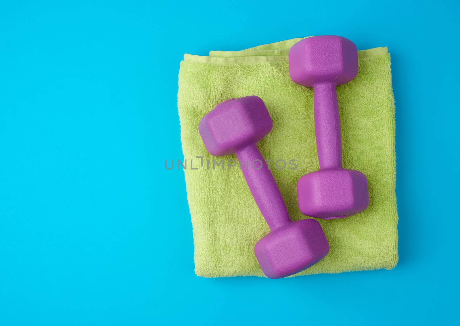 pair of lilac plastic dumbbells lie on a light green terry towel, blue background from a rug, backdrop for sports, copy space