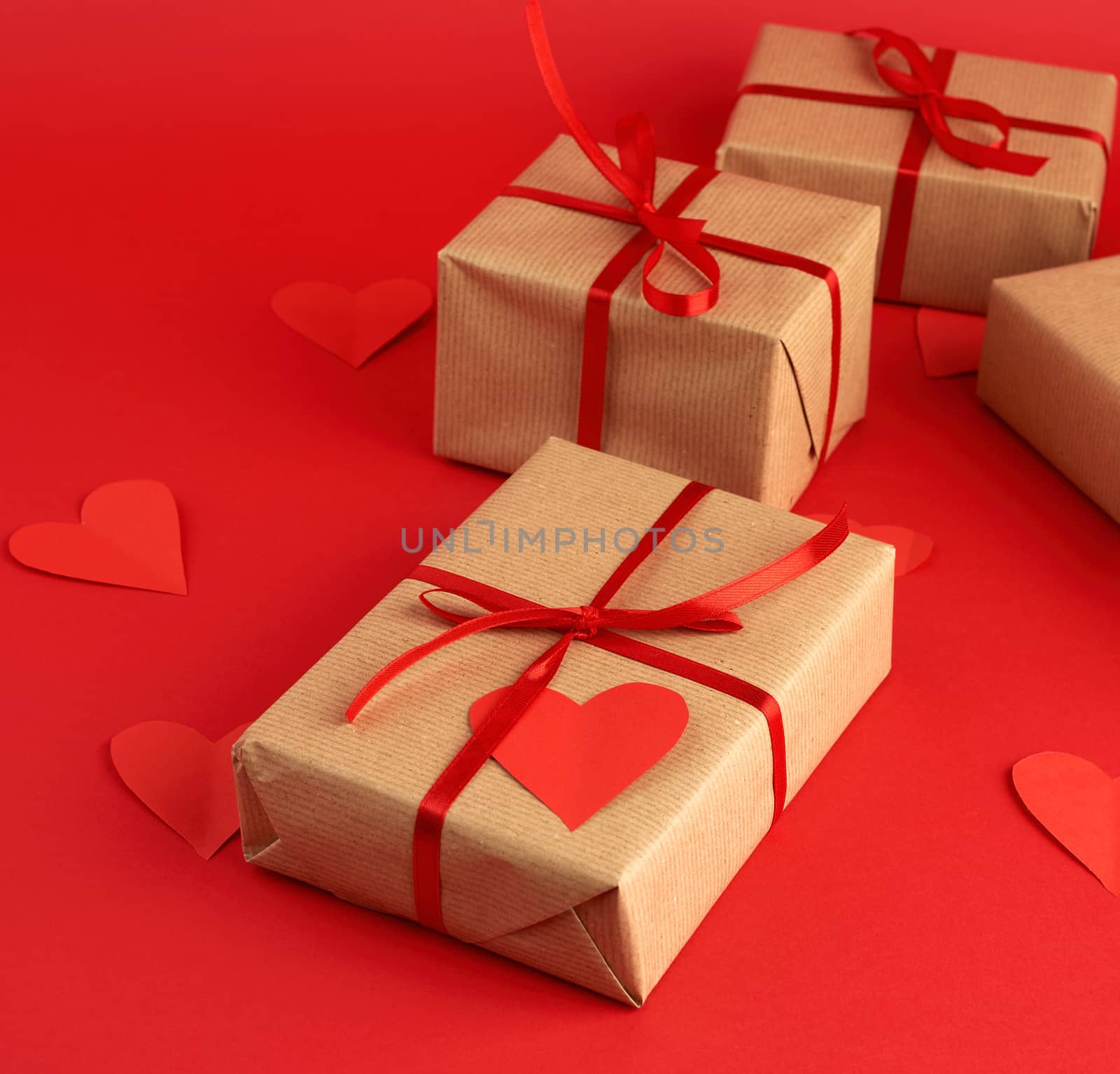 box wrapped in brown kraft paper and tied with a red thin silk ribbon, cut out of paper hearts, red background, gift for Valentine's Day, birthday, flat lay