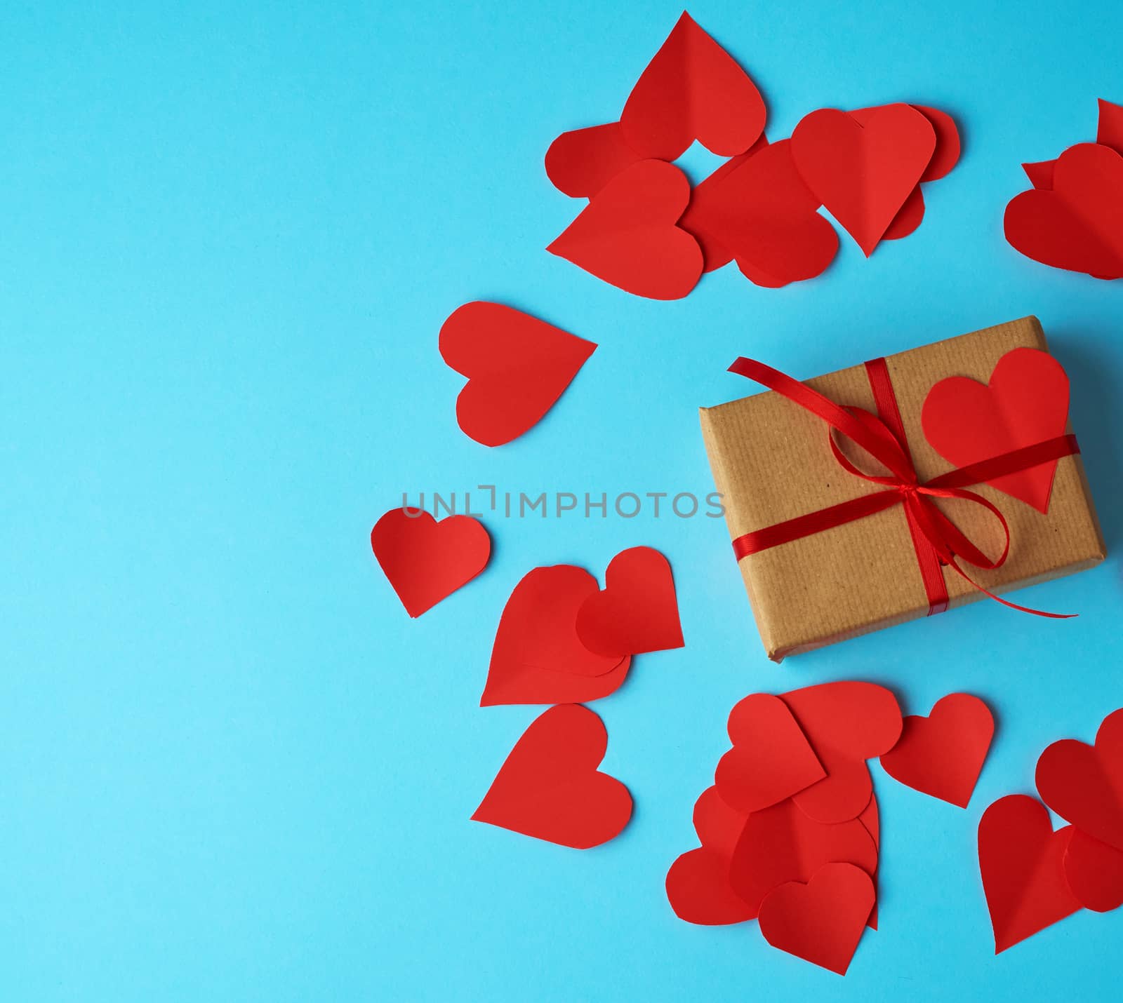 rectangular box is wrapped in brown paper and tied with a red thin silk ribbon on a blue background, red cut out paper hearts, copy space, holiday is Valentine's Day