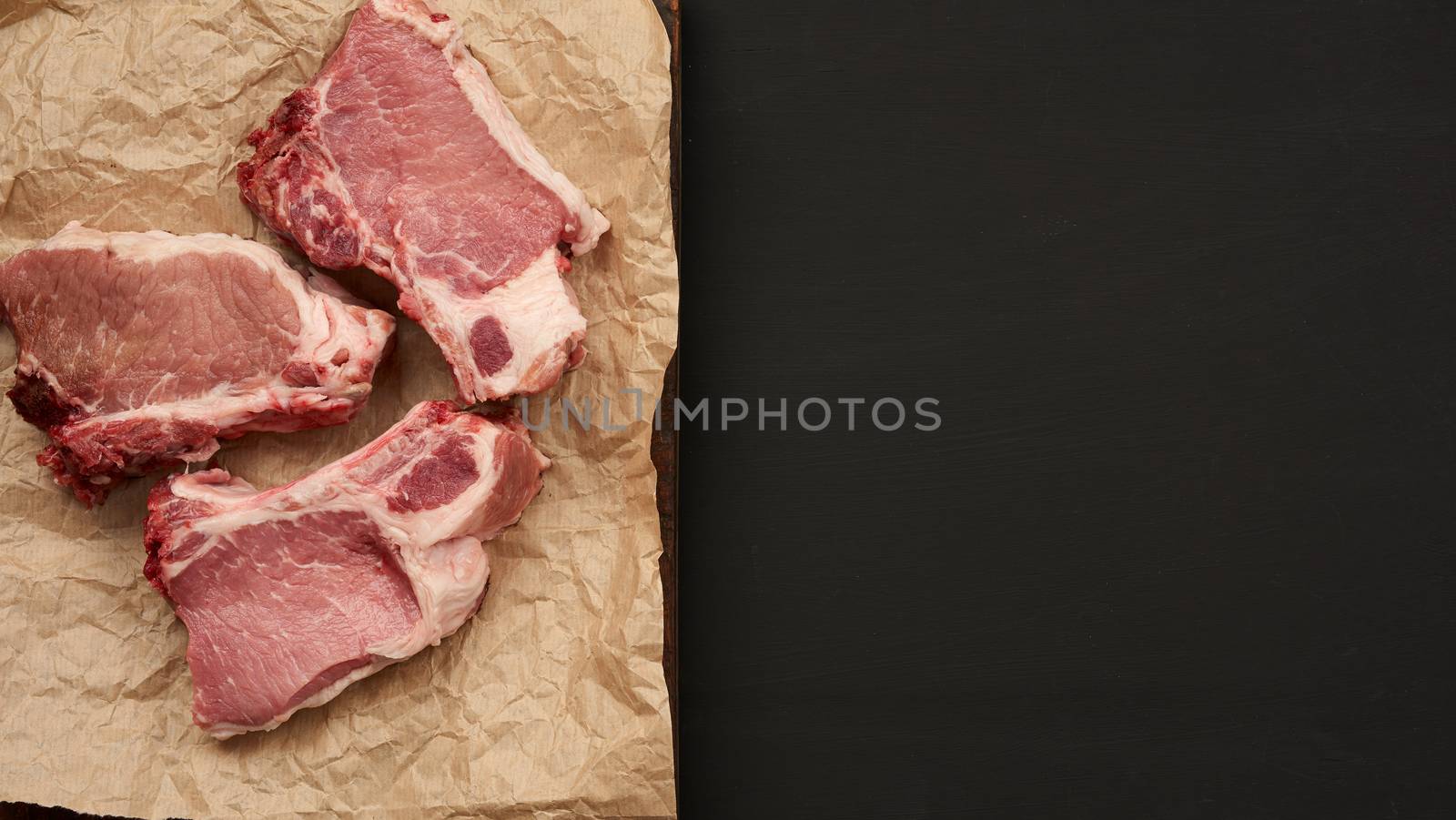 raw juicy pork slices of meat on the rib, food lies on brown parchment paper,  black wooden background and an empty space for writing a recipe or menu, top view