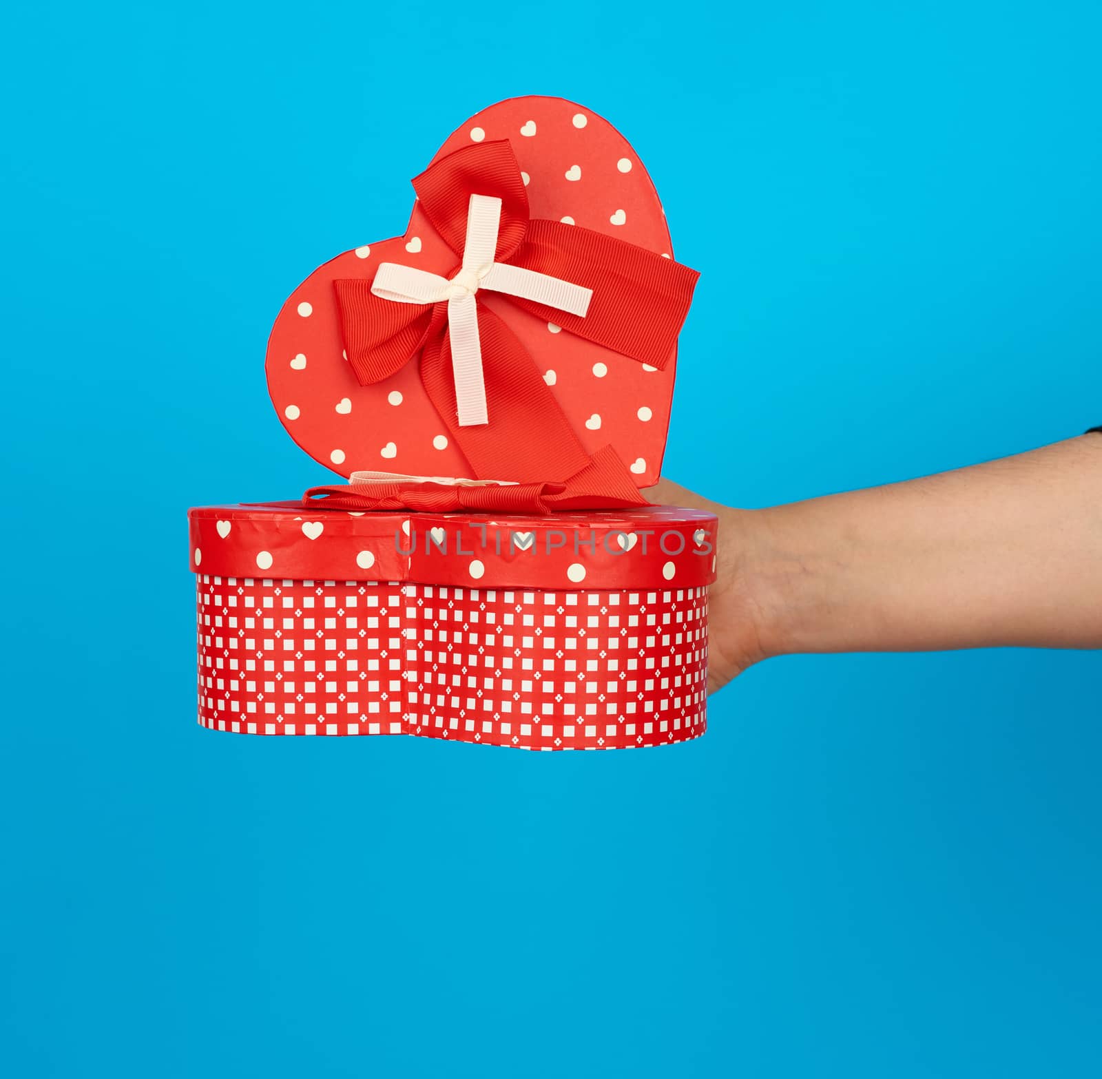 hand holds a red heart-shaped cardboard box with silk ribbon on a blue background, gift giving concept