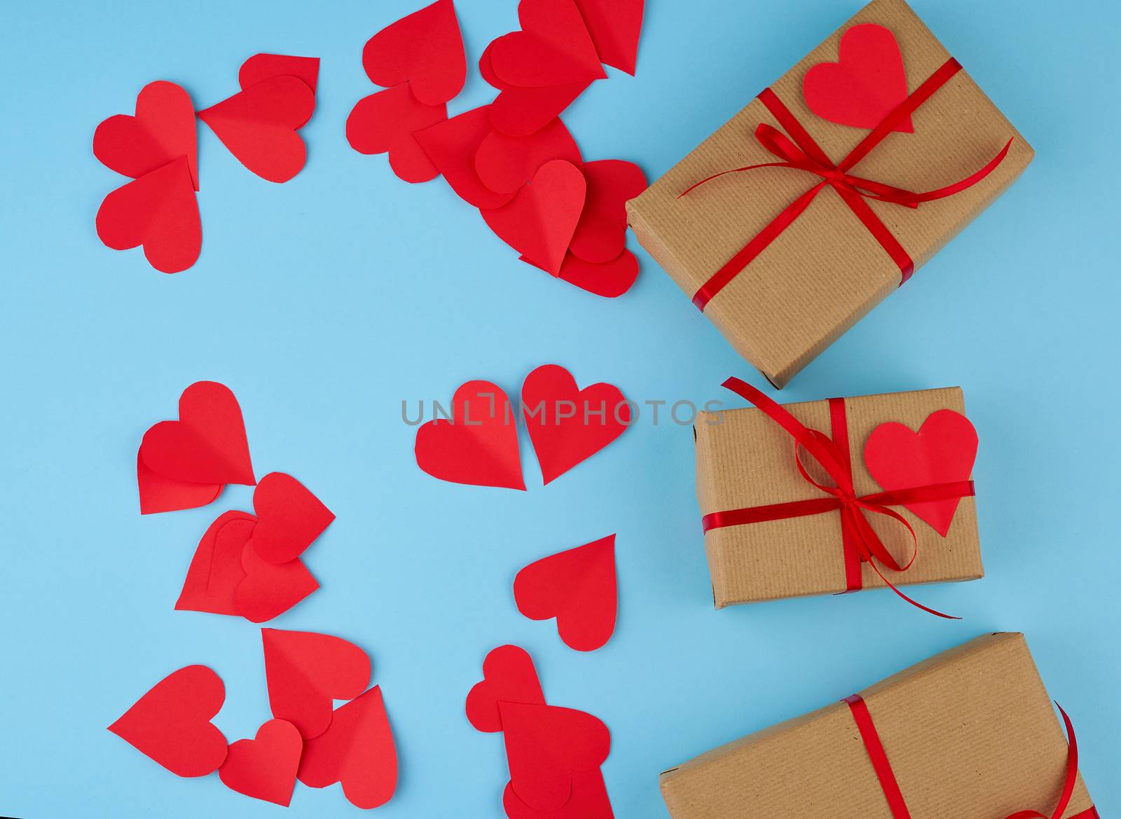 rectangular box is wrapped in brown paper and tied with a red thin silk ribbon on a blue background, red cut out paper hearts, copy space, holiday is Valentine's Day
