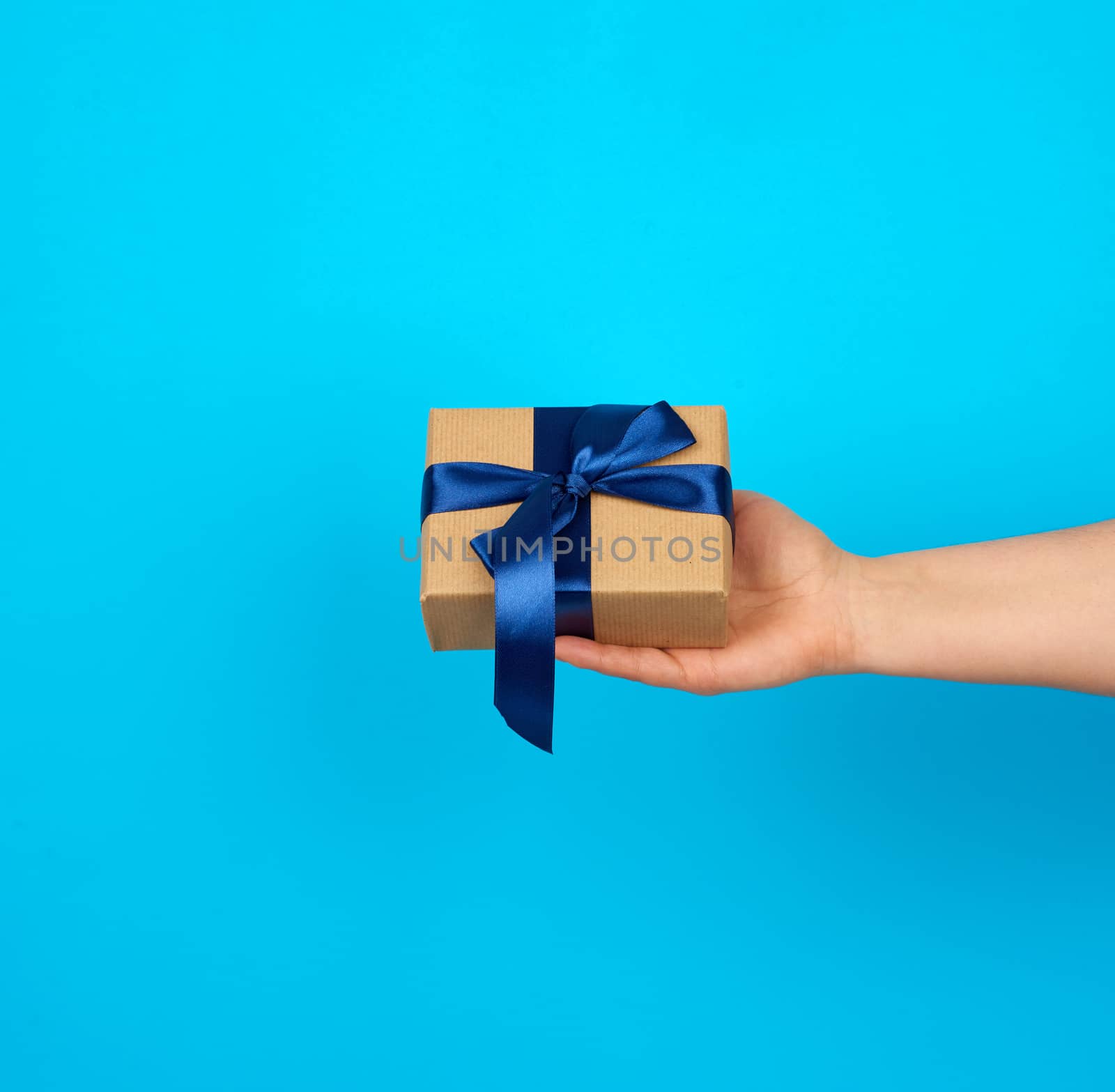 hand holds a box with a gift wrapped in brown kraft paper and tied with a silk blue ribbon on a blue background, concept of giving a gift