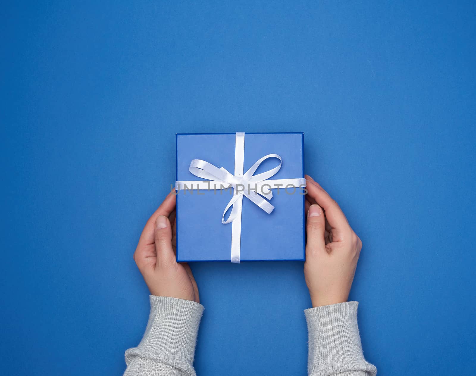 two female hands in a sweater hold a square blue box with a bow on a blue classic background, top view, festive backdrop