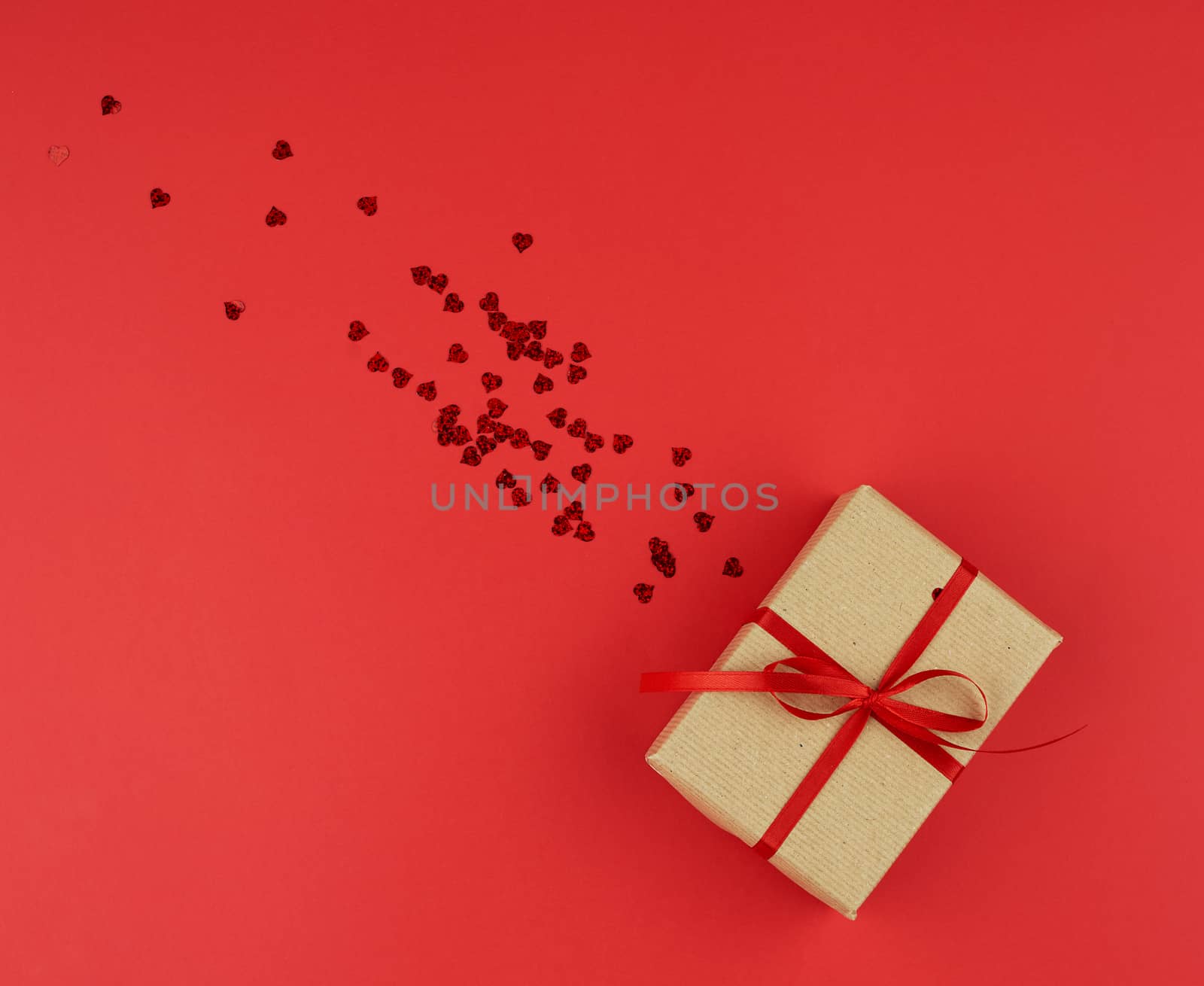 box wrapped in brown kraft paper and tied with a red thin silk ribbon, scattered confetti in the shape of hearts, red background, festive backdrop for Valentine's Day