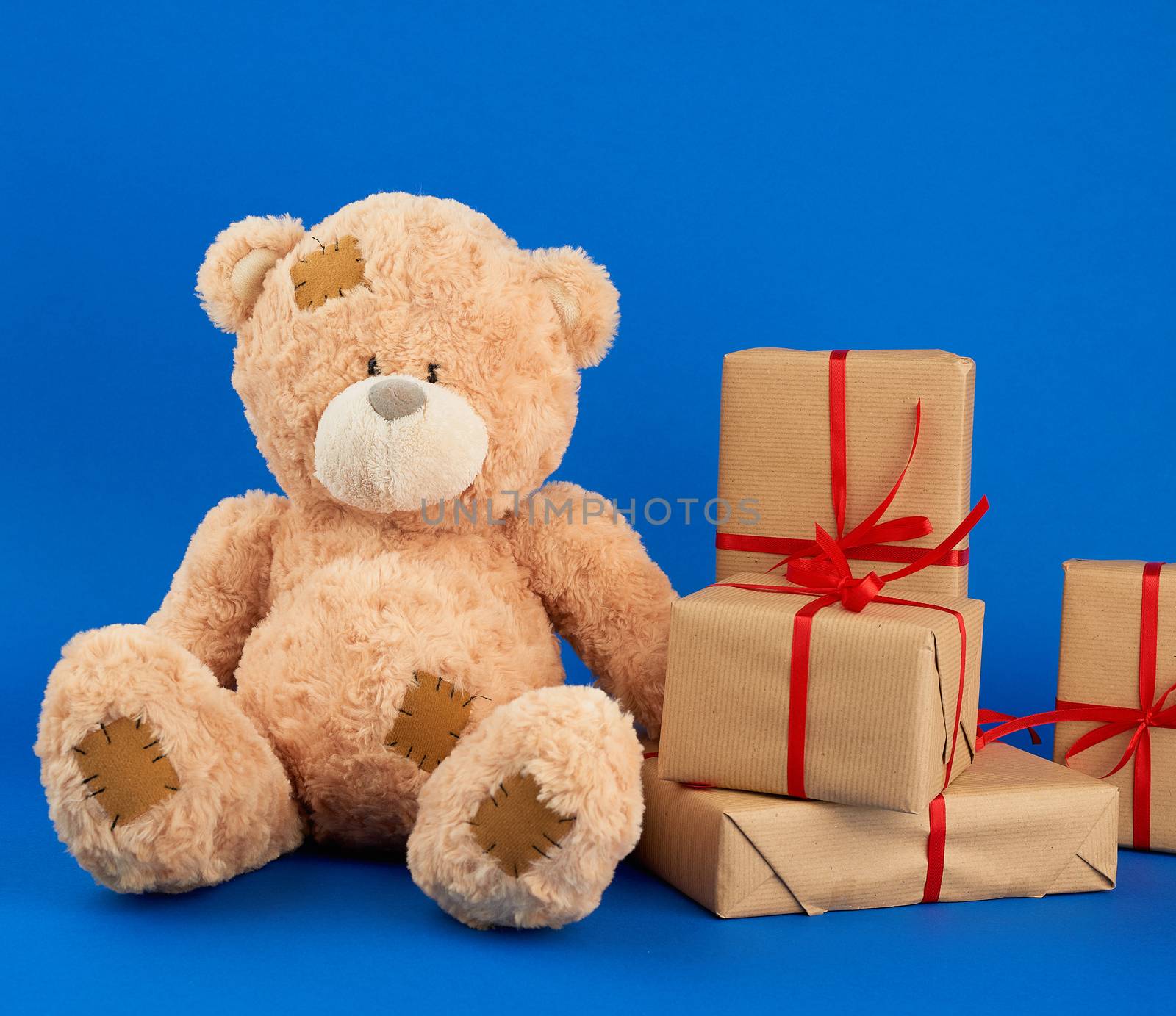 big beige teddy bear with patches, stack of gifts in boxes wrapped in brown paper, tied with silk ribbon on a blue background, happy birthday and valentines day